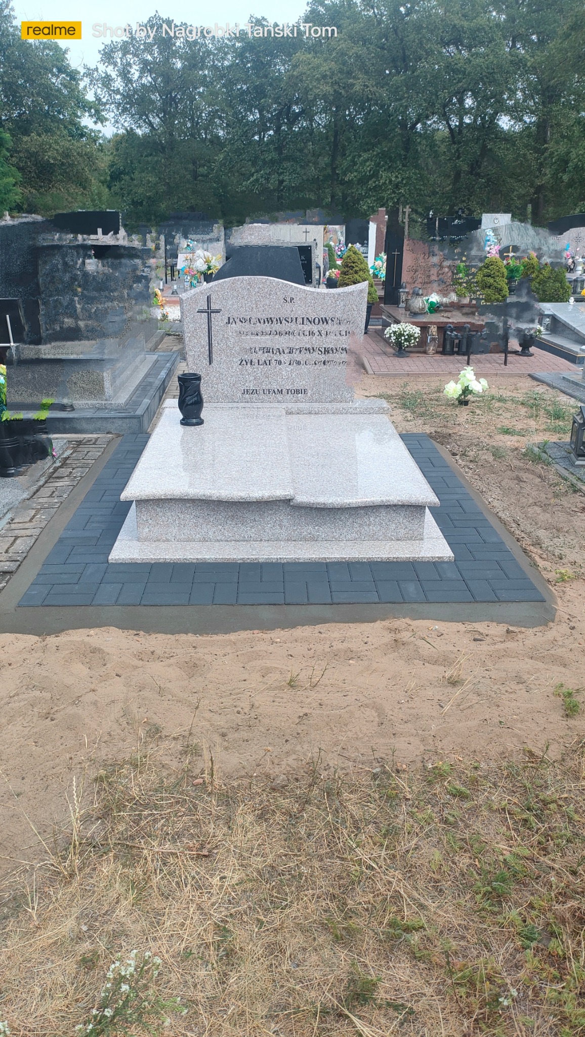 Jasny, granitowy nagrobek z krzyżem i czarnym wazonem, otoczony szarą kostką brukową na cmentarzu. Widoczne inne nagrobki i zieleń w tle.