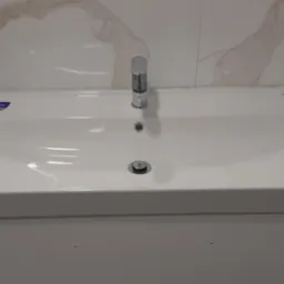 Biała umywalka Cersanit z chromowaną baterią, zamontowana na szafce w łazience wykończonej płytkami imitującymi marmur, z widocznym włącznikiem światła.