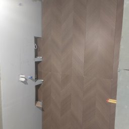 Krzysztof Danowski - Ściana łazienki z ułożonymi brązowymi płytkami w jodełkę. Widoczne wnęki z półkami i przygotowane przyłącza wodne. Ściana w trakcie prac wykończeniowych.