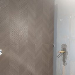 Krzysztof Danowski - Ściana łazienki z ułożonymi płytkami w jodełkę, obok ściana przygotowana pod instalację hydrauliczną z wystającymi rurami. Widoczny etap prac wykończeniowych.