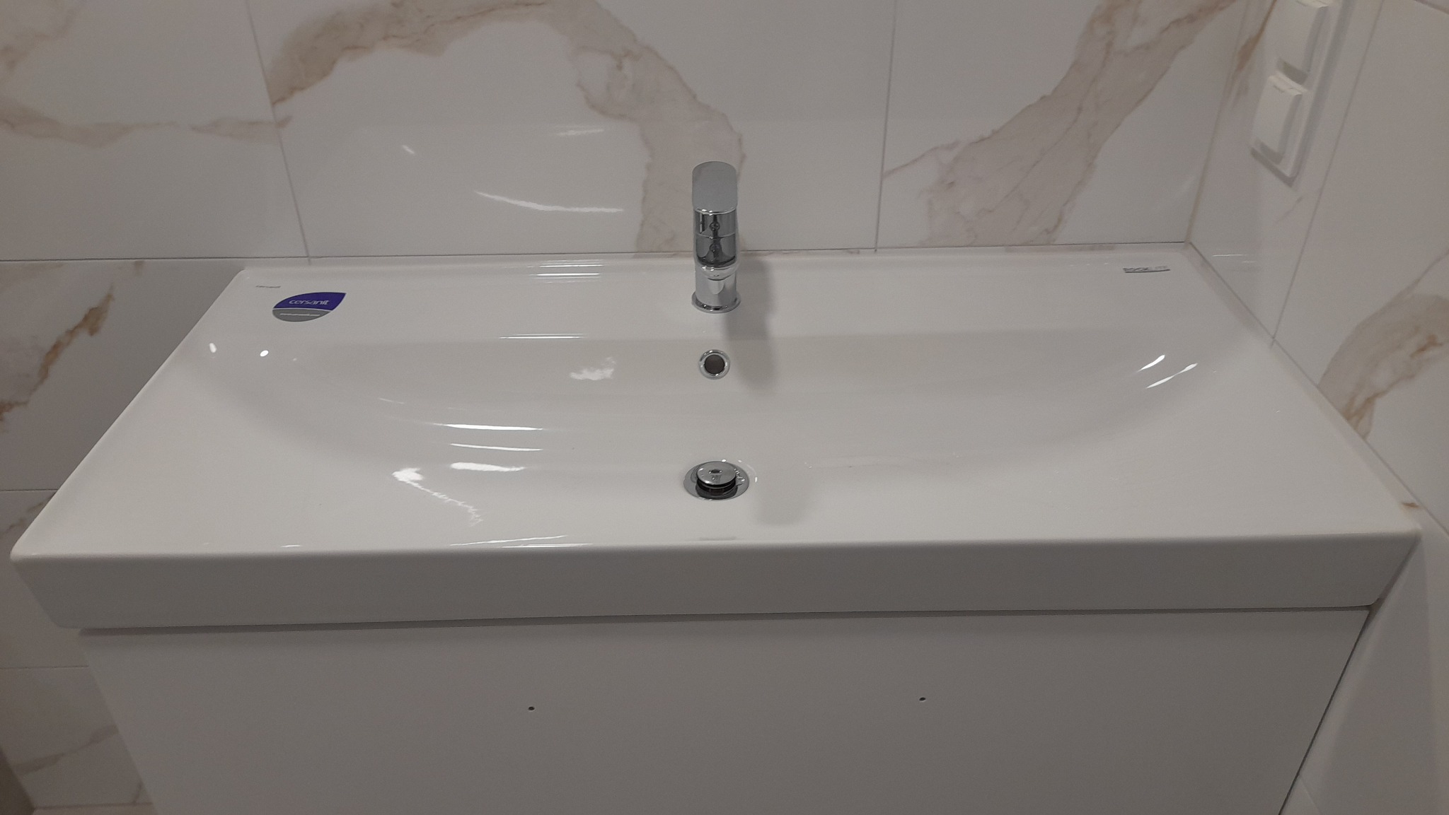 Biała umywalka Cersanit z chromowaną baterią, zamontowana na szafce w łazience wykończonej płytkami imitującymi marmur, z widocznym włącznikiem światła.