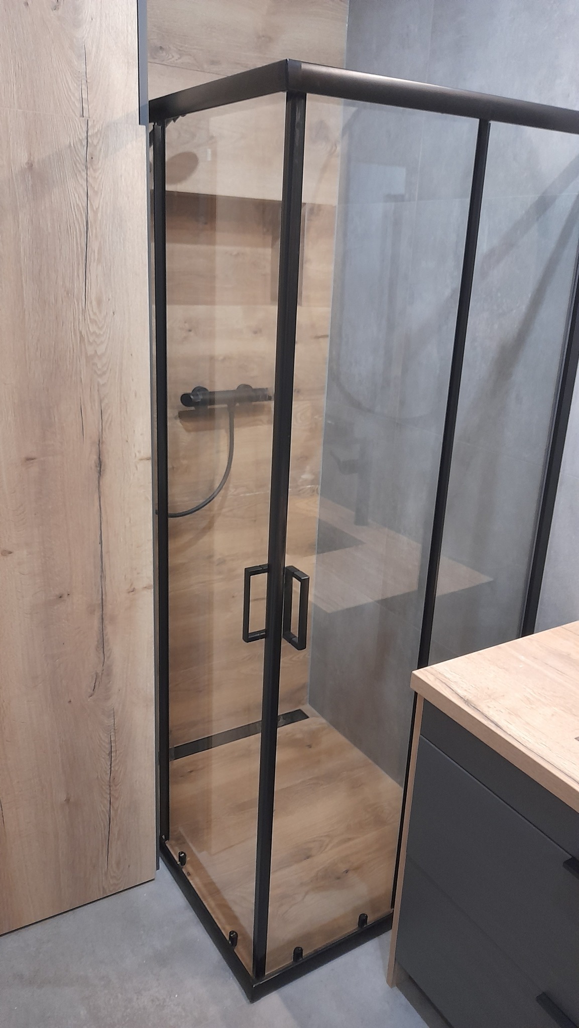 Kabina prysznicowa z czarną ramą, imitująca drewno podłoga i ściana, szara ściana w tle, nowoczesna aranżacja łazienki.