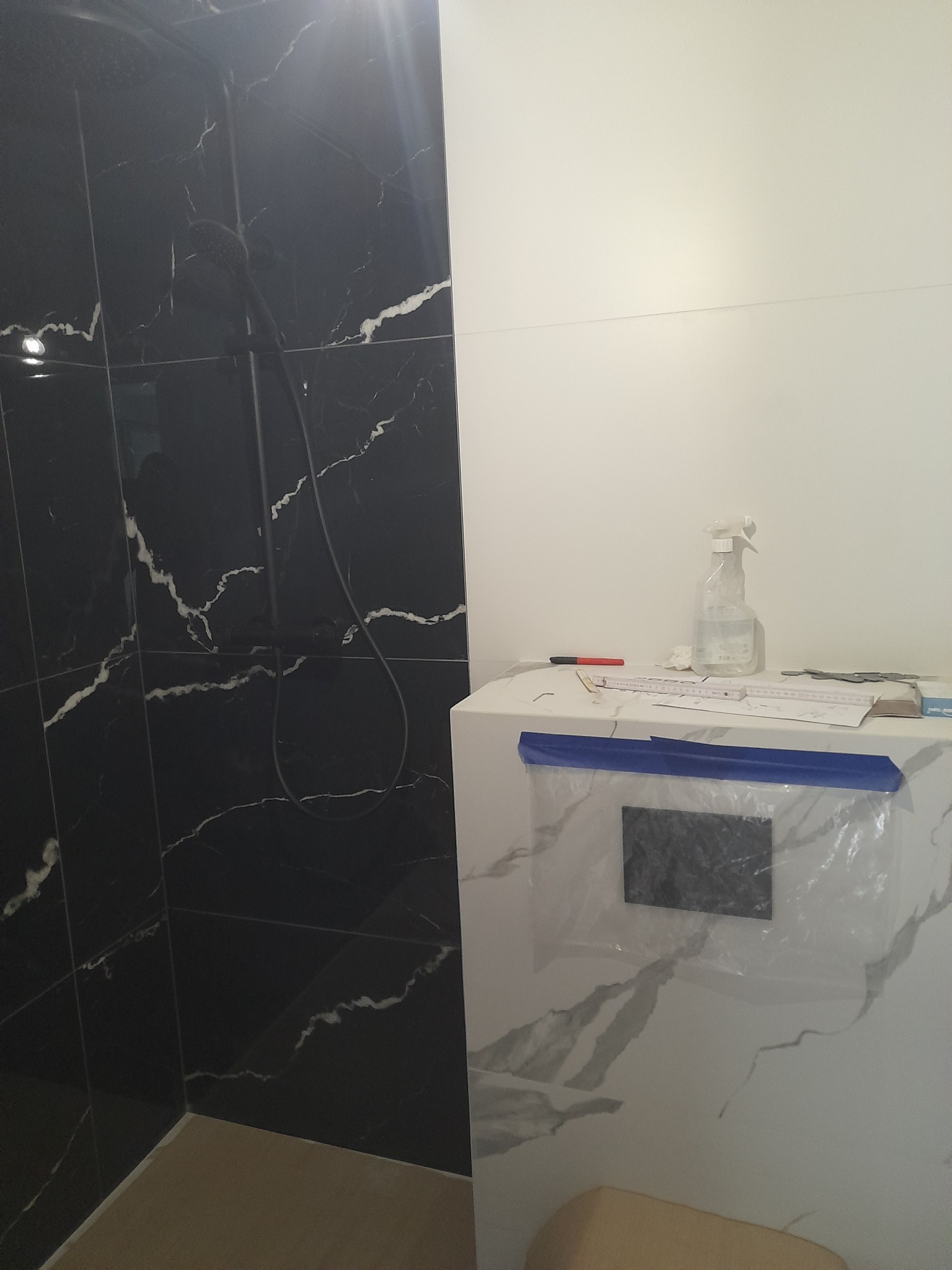 Wykończona kabina prysznicowa z czarnymi płytkami imitującymi marmur, biała zabudowa z płytkami, zabezpieczona folią i taśmą malarską, widoczne narzędzia i butelka ze spryskiwaczem.