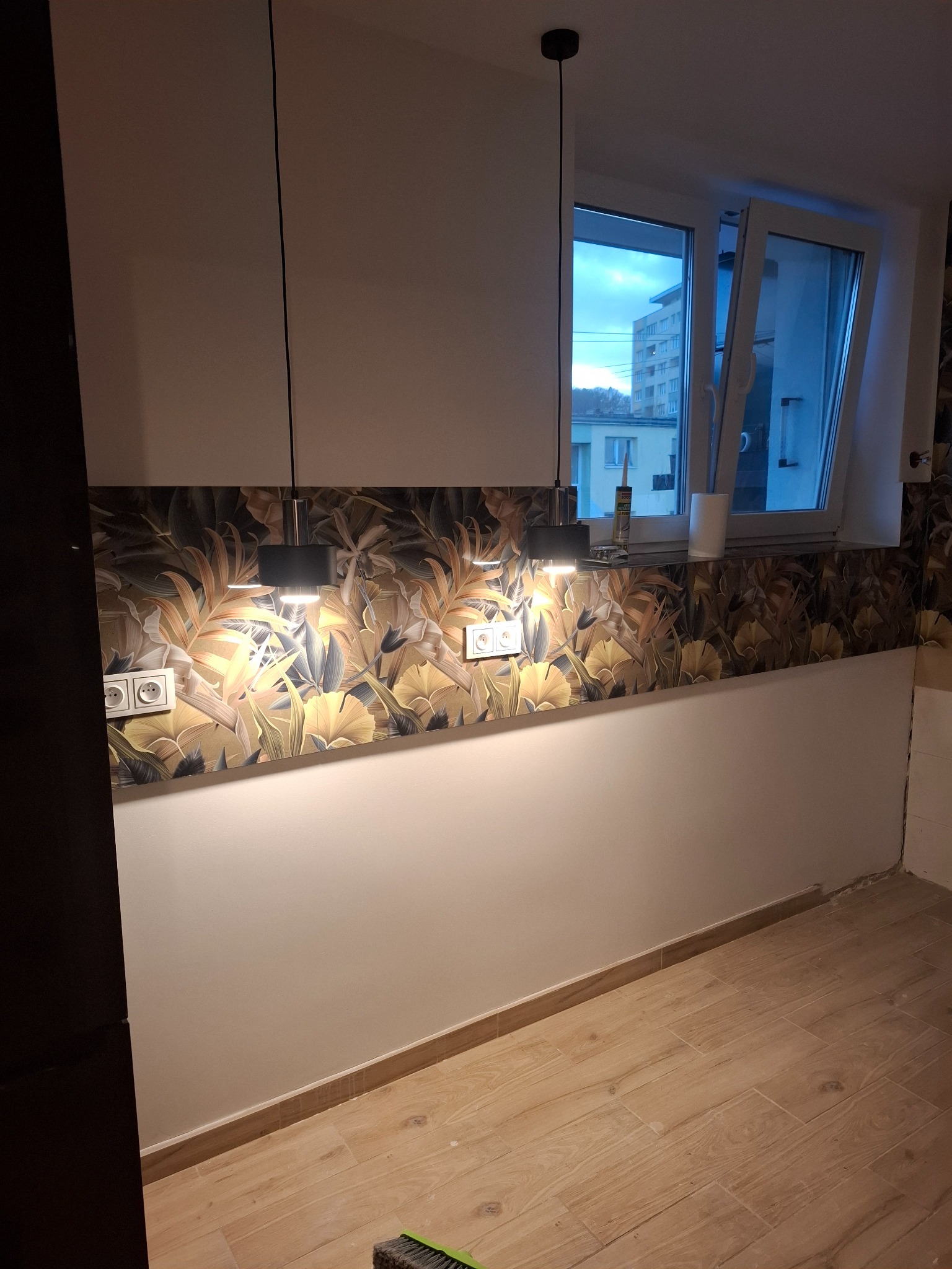 Wykończona ściana z wzorzystą tapetą w tropikalne liście, oświetlona dwoma wiszącymi lampami. Widoczne gniazdka elektryczne i fragment okna z widokiem na budynek.