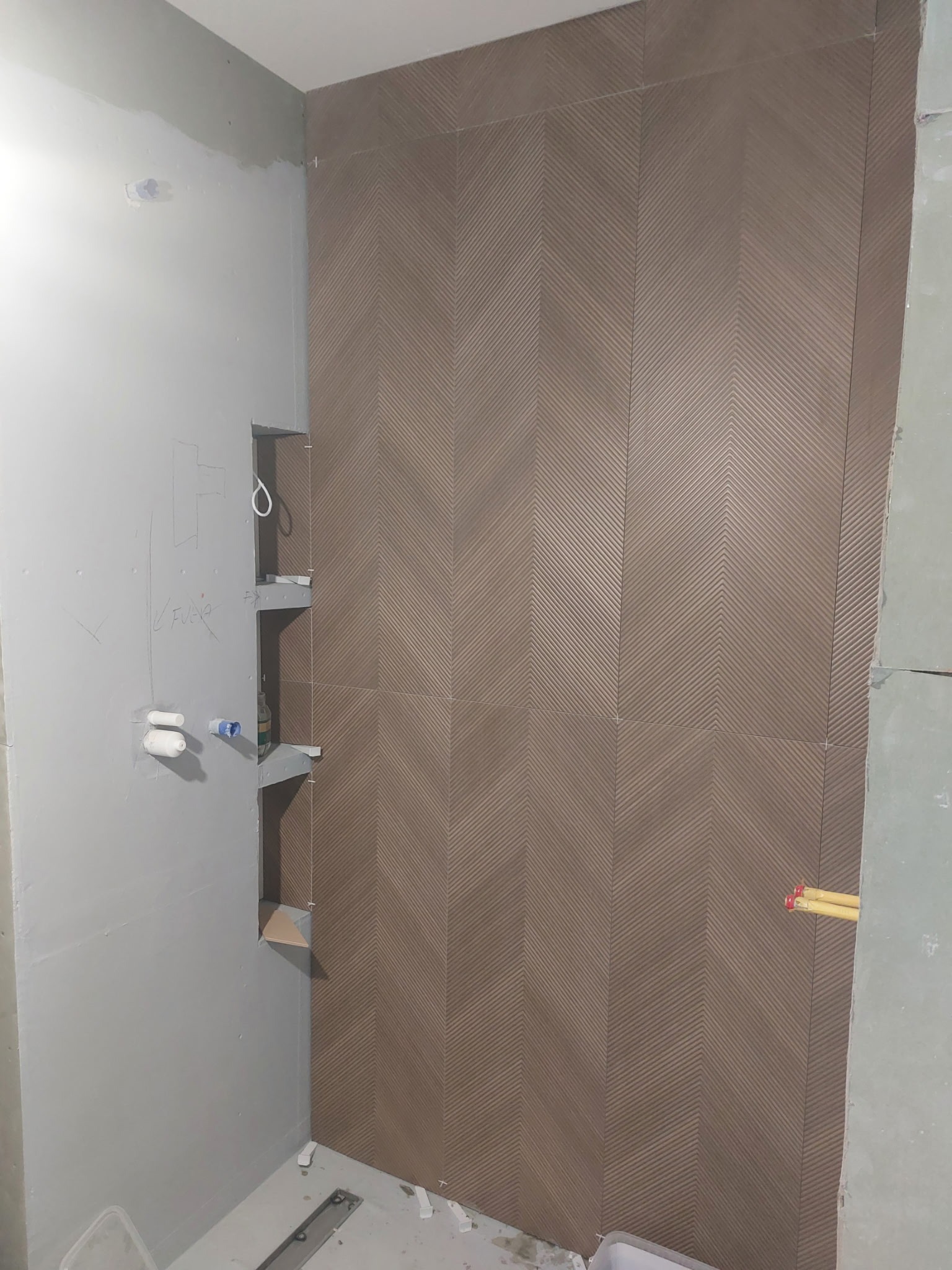 Ściana łazienki z ułożonymi brązowymi płytkami w jodełkę. Widoczne wnęki z półkami i przygotowane przyłącza wodne. Ściana w trakcie prac wykończeniowych.