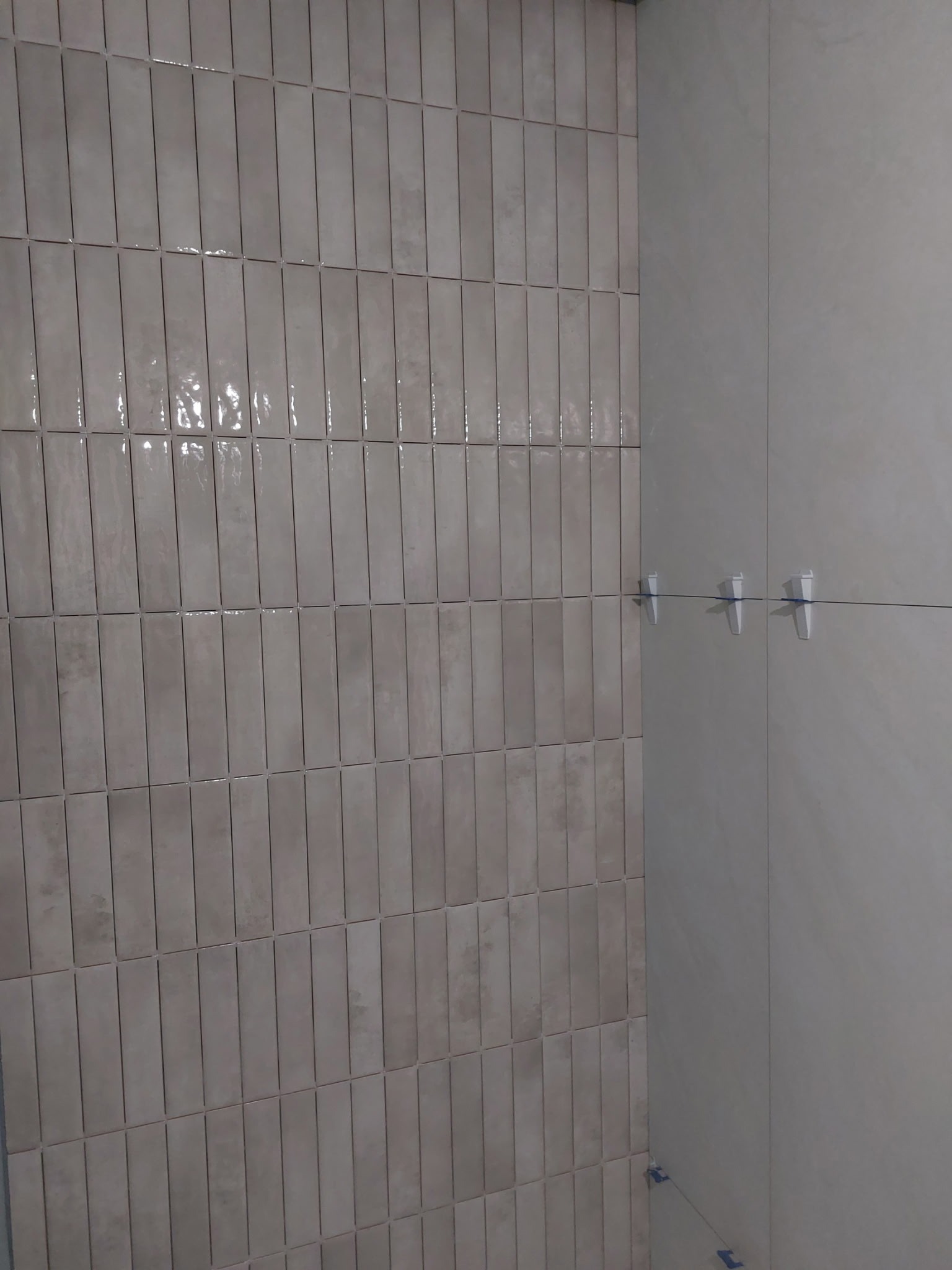 Ułożone pionowo, beżowe, błyszczące płytki w kształcie prostokątów na ścianie. Widoczne krzyżyki dystansowe. Po prawej gładka, jasna ściana.