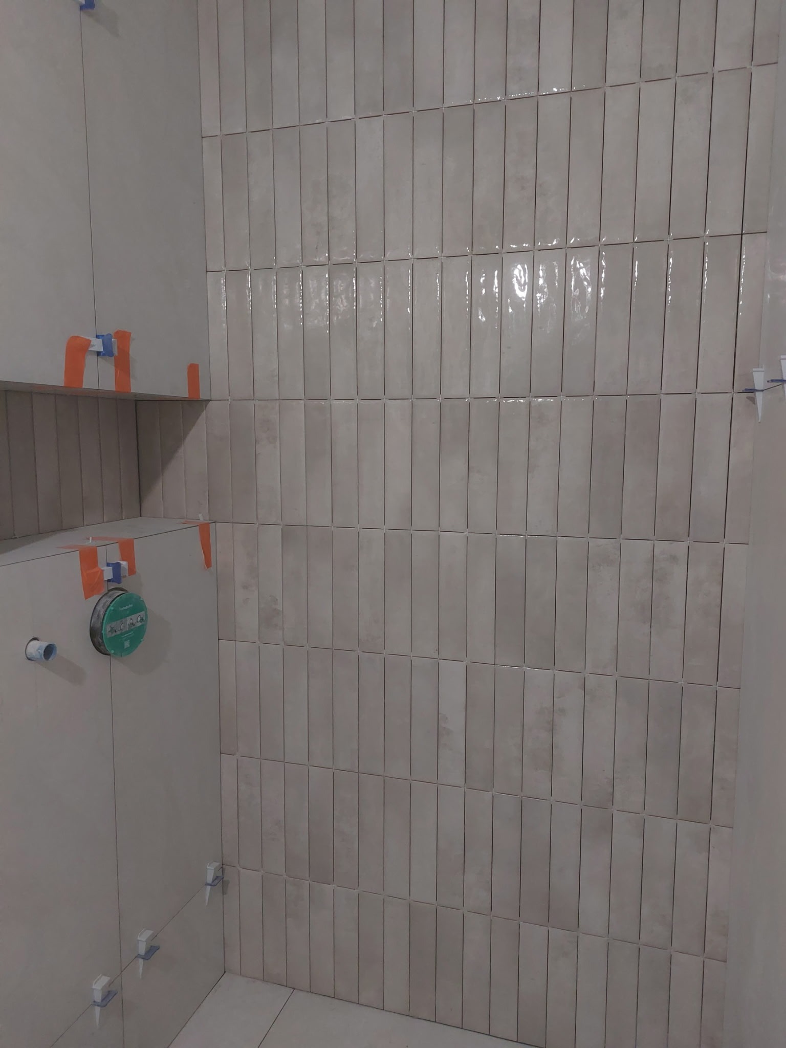 Ściana w łazience wyłożona błyszczącymi, beżowymi płytkami w kształcie wąskich prostokątów, z widocznymi krzyżykami dystansowymi i fragmentem półki.