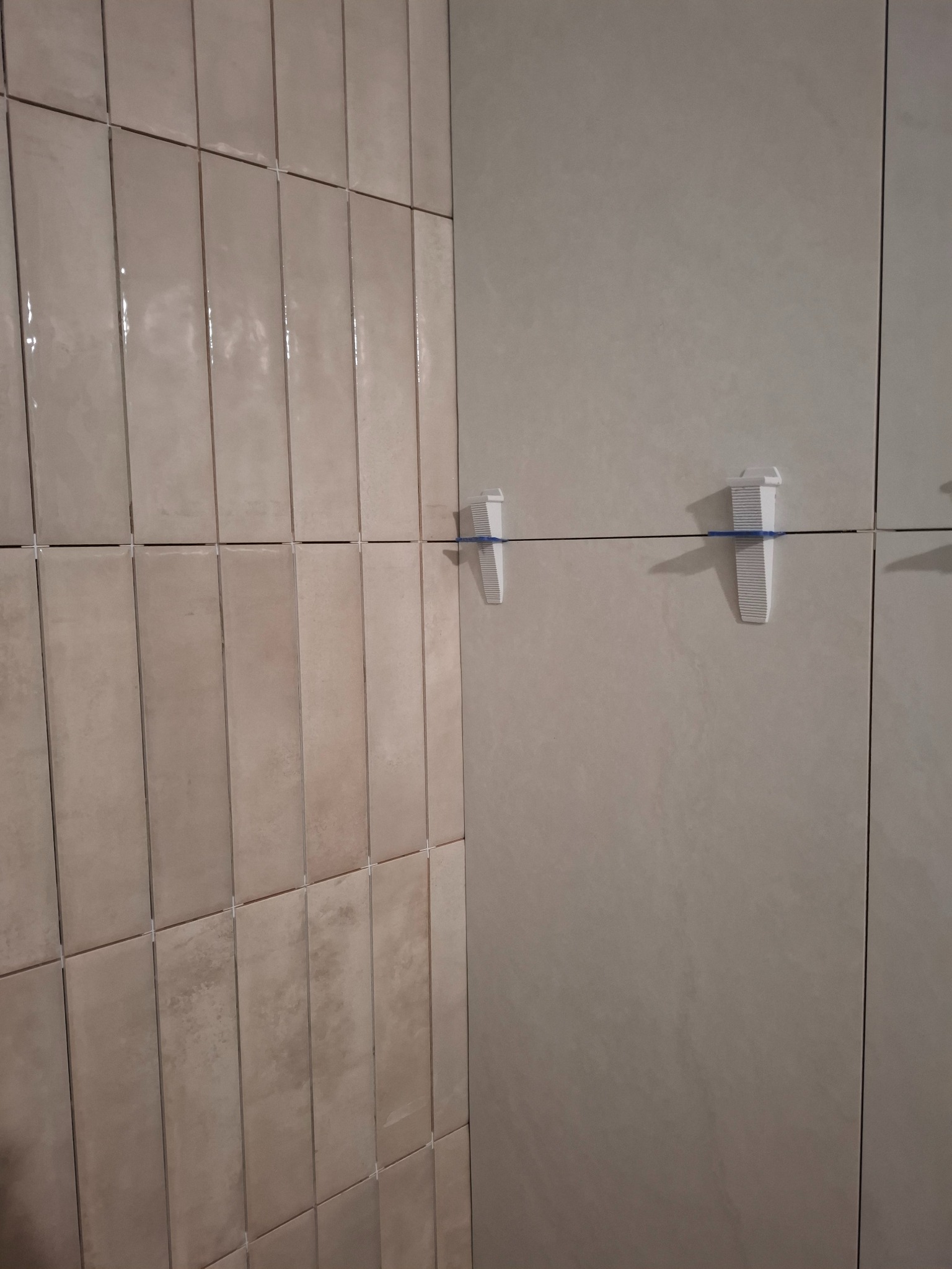 Układanie beżowych, wąskich płytek na ścianie z użyciem systemu poziomowania. Widoczne krzyżyki dystansowe i kliny. Częściowo ułożona ściana.