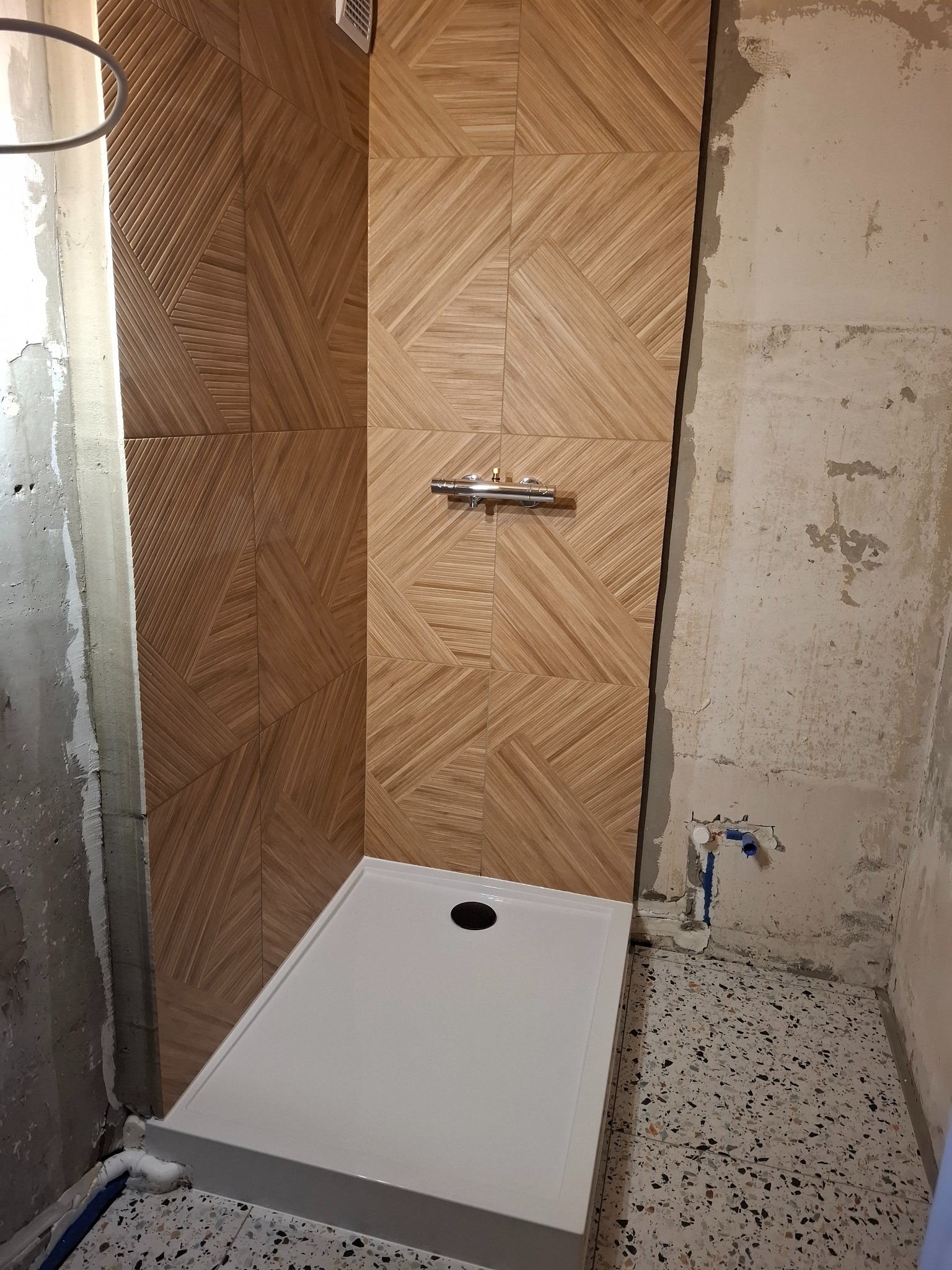Montaż kabiny prysznicowej z drewnopodobnymi płytkami w geometryczny wzór. Widoczne surowe ściany i podłoga w trakcie prac wykończeniowych.