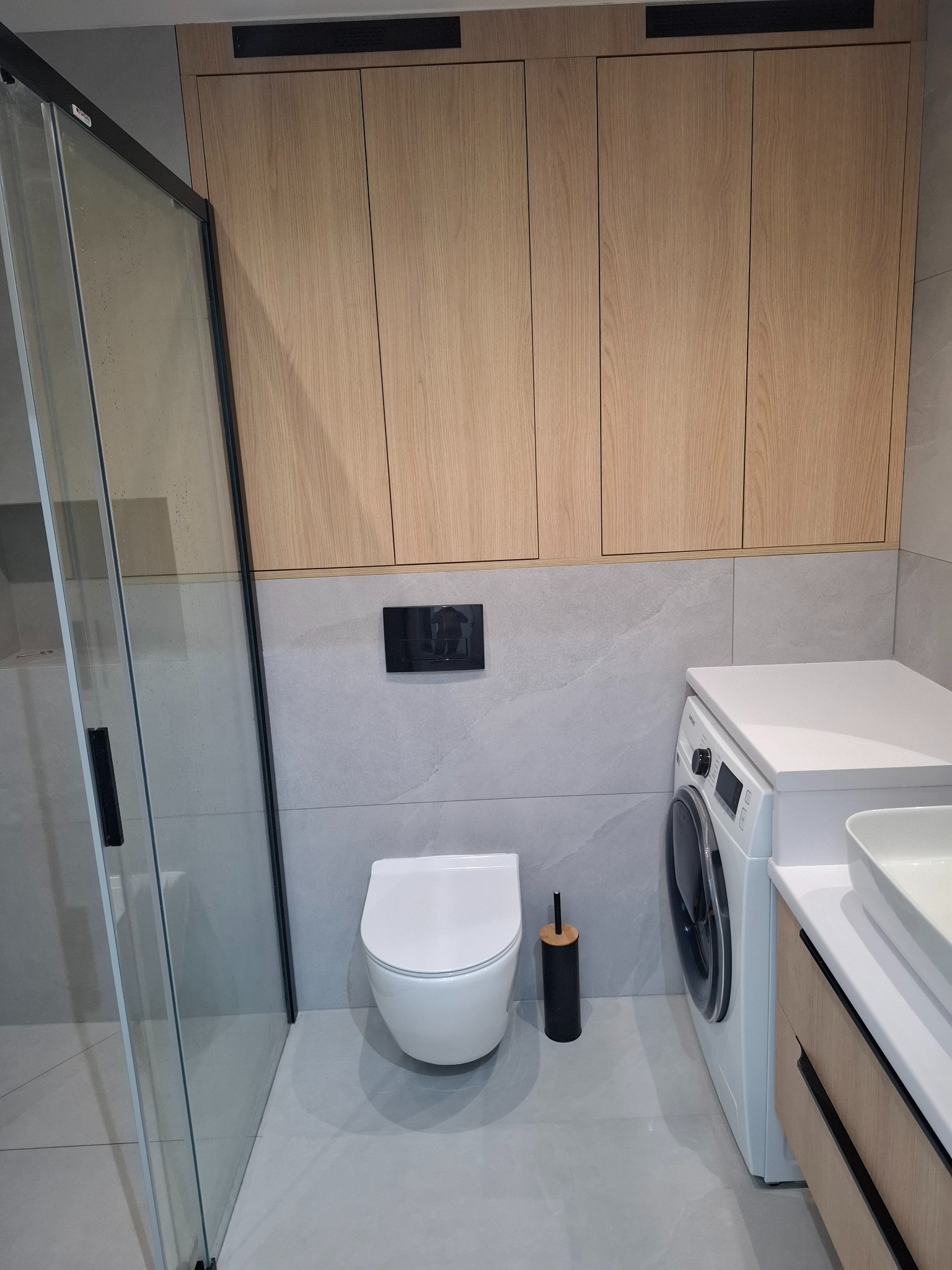 Jasna łazienka po remoncie: kabina prysznicowa z czarnymi profilami, podwieszana toaleta, pralka z umywalką na blacie i szafki wiszące z drewnianymi frontami.