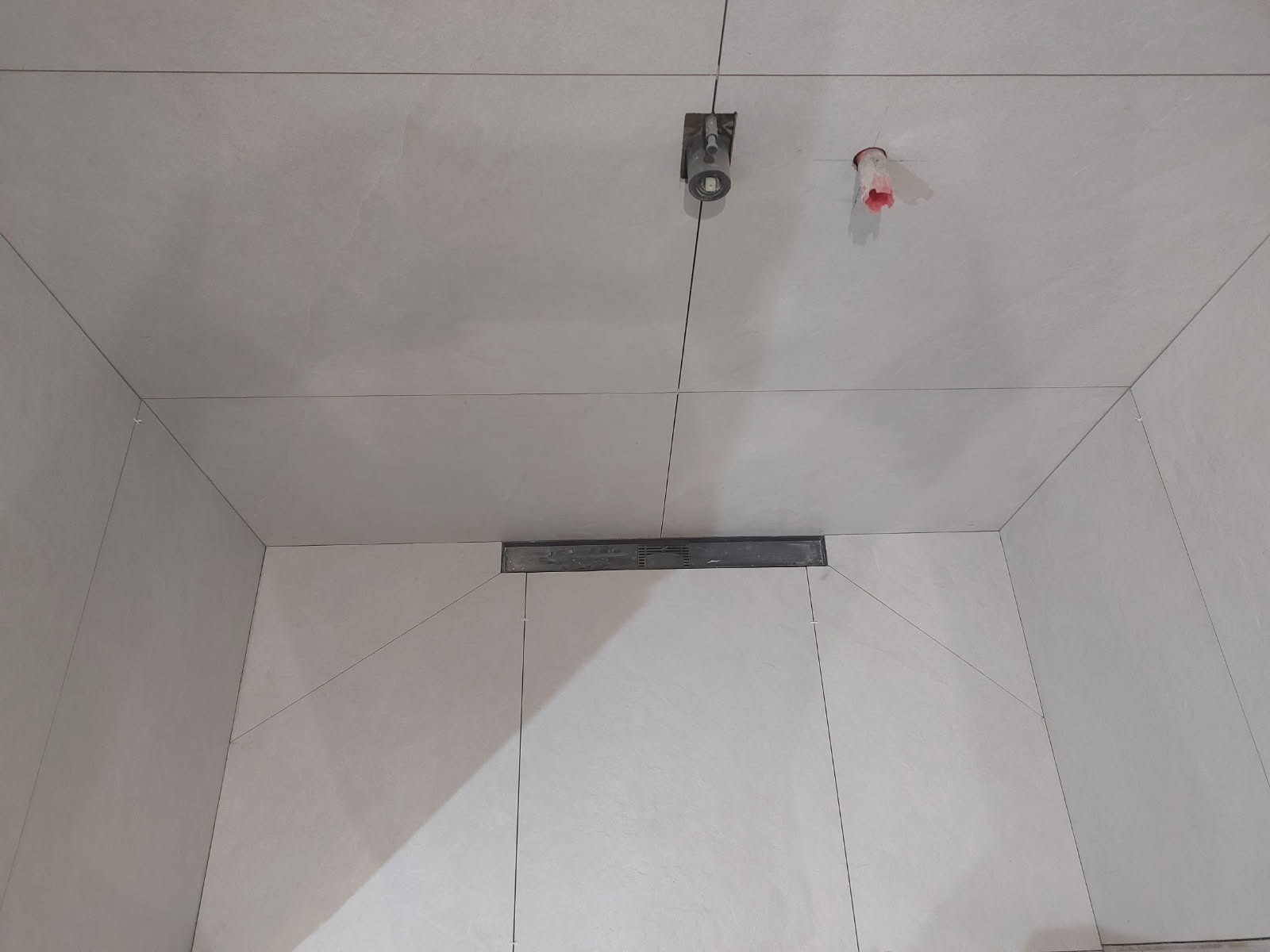 Wykończona łazienka z szarymi płytkami, widoczny odpływ liniowy, punktowe oświetlenie i fragment instalacji. Minimalistyczny design, surowe wykończenie.