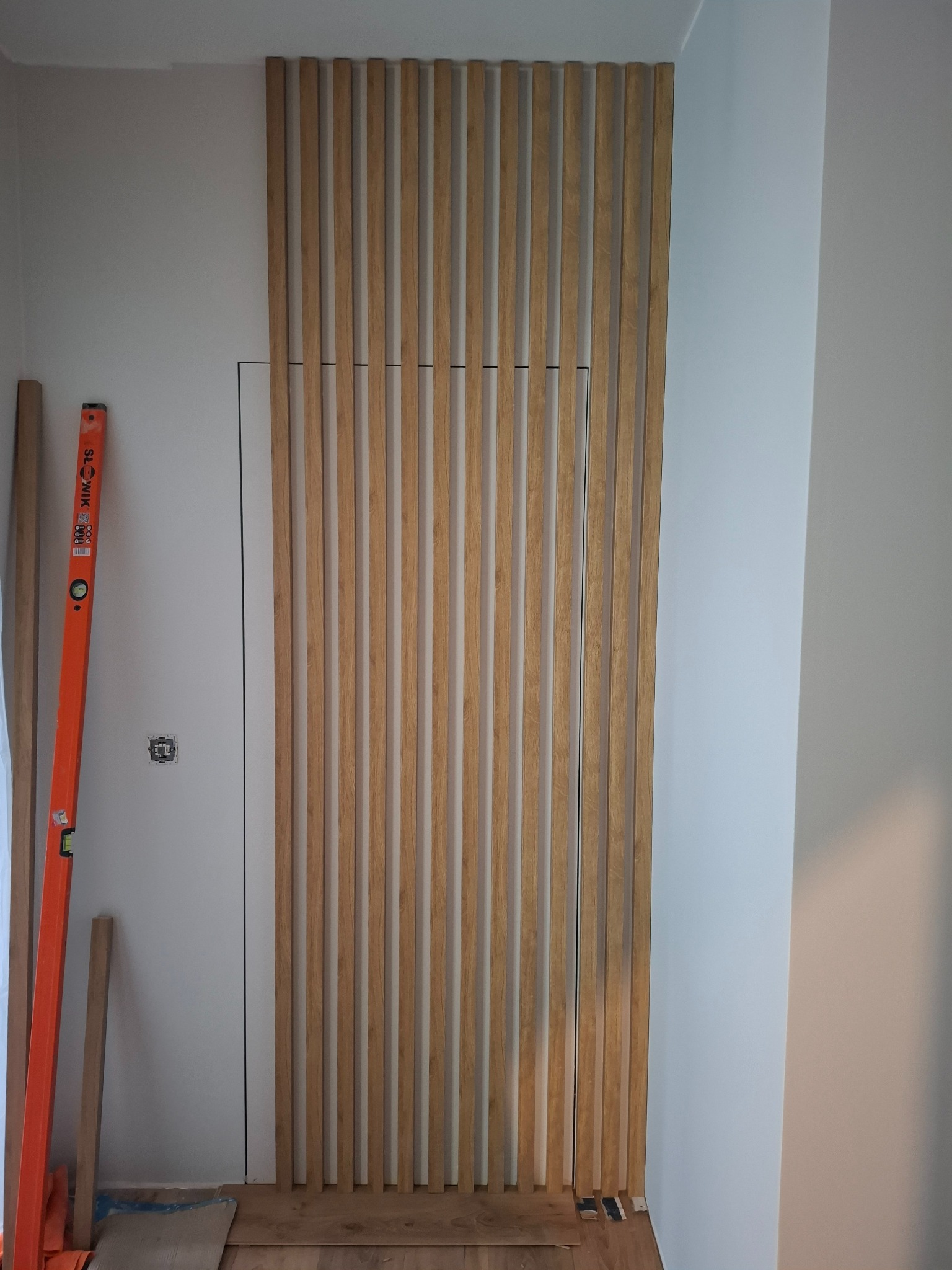 Drewniane lamele na ścianie, ukrywające drzwi, z poziomnicą i fragmentem instalacji elektrycznej. Wykończenie wnętrz w trakcie realizacji.