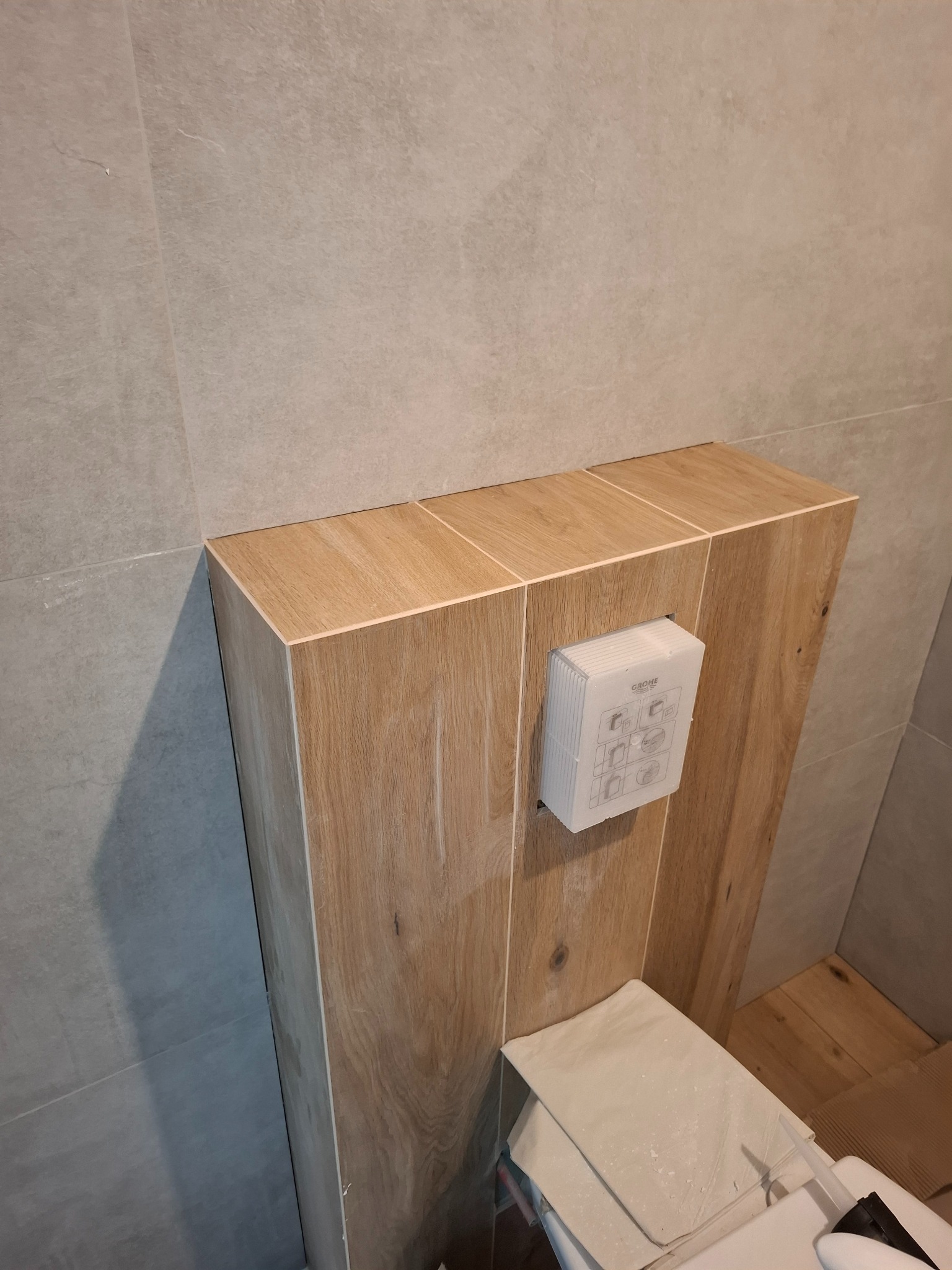 Obudowa stelaża WC z drewnopodobnych płytek, szare płytki ścienne, biały przycisk spłukujący Grohe. Ukończona zabudowa w łazience.