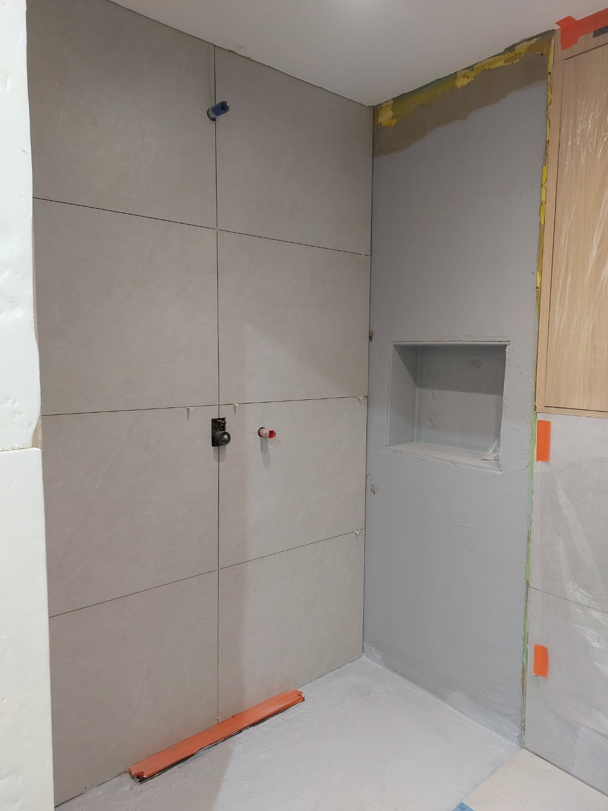 Ściana łazienki w trakcie wykańczania: ułożone jasne płytki, widoczne elementy instalacji, wnęka w ścianie. Fragment zabezpieczonej szafki.