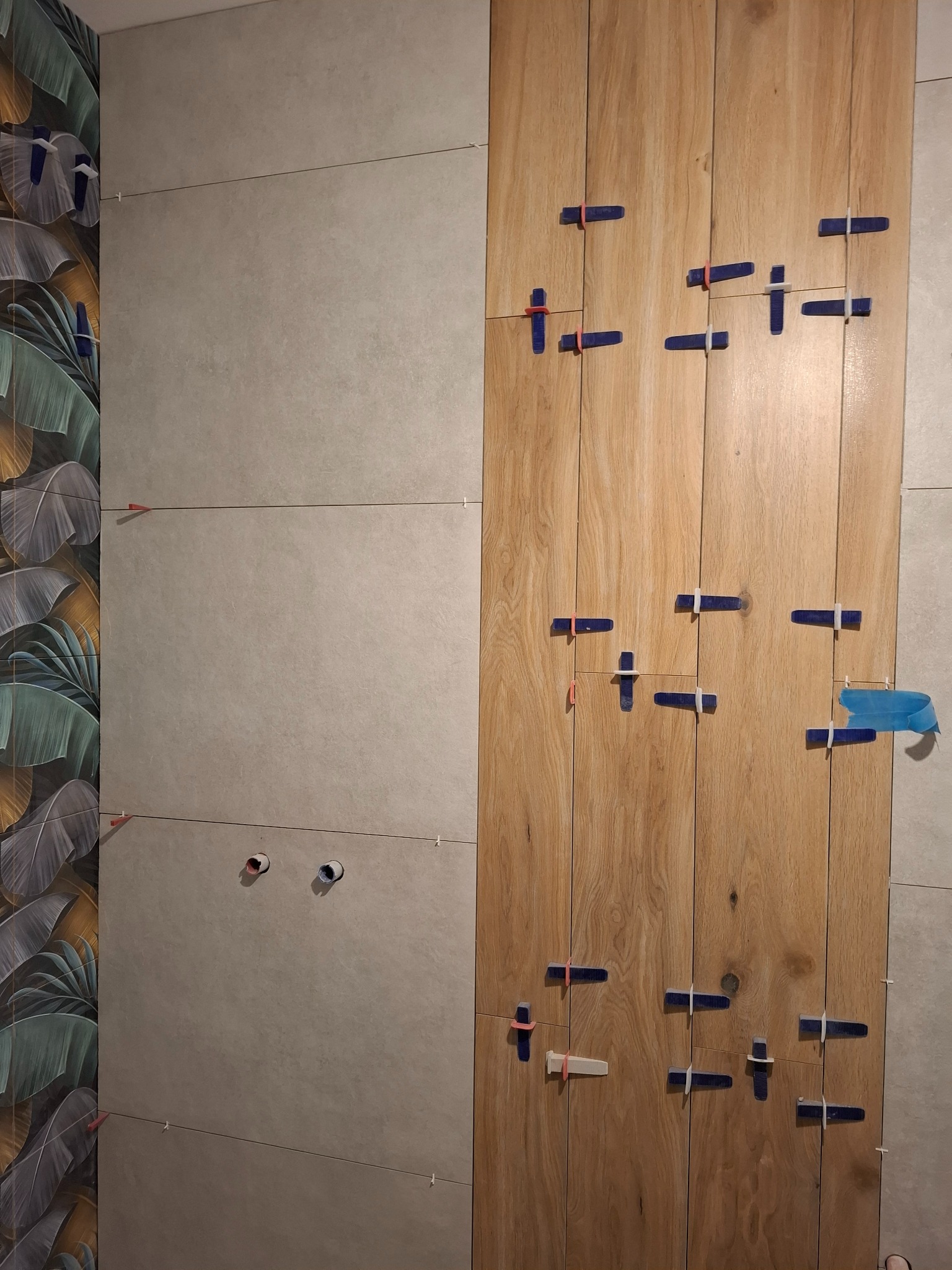 Układanie płytek imitujących drewno na ścianie, z użyciem klipsów poziomujących. Widoczne otwory na instalacje i fragment ściany z wzorem liści.