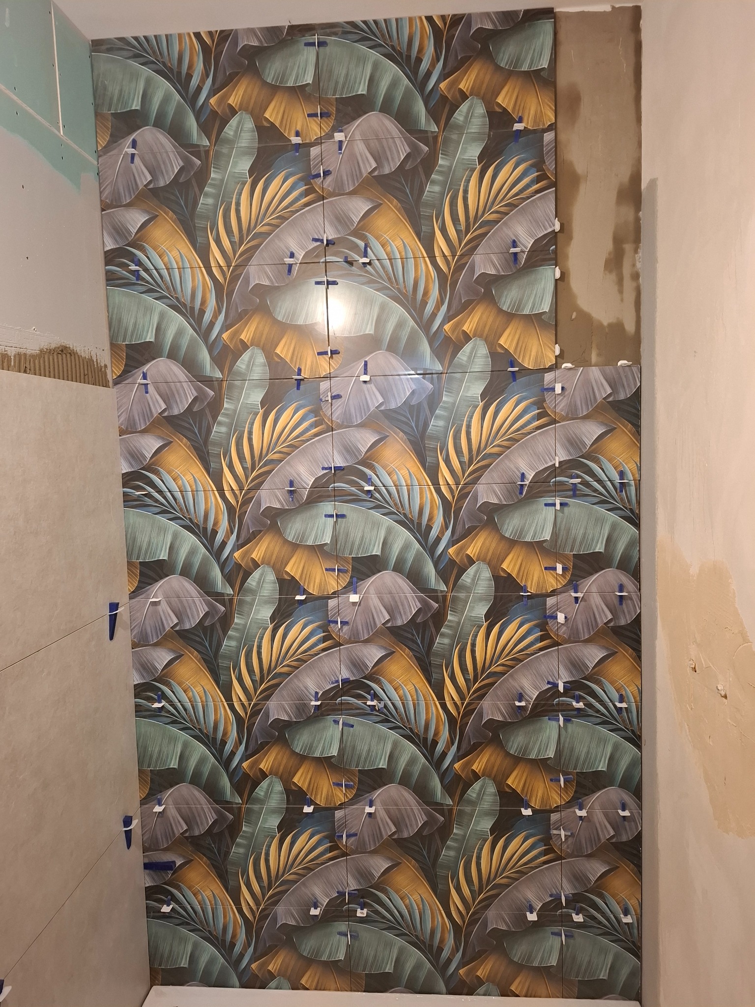 Układanie płytek z motywem liści na ścianie. Widoczne krzyżyki dystansowe. Częściowo wykończona ściana z płytkami, obok surowa ściana przygotowana do dalszych prac.