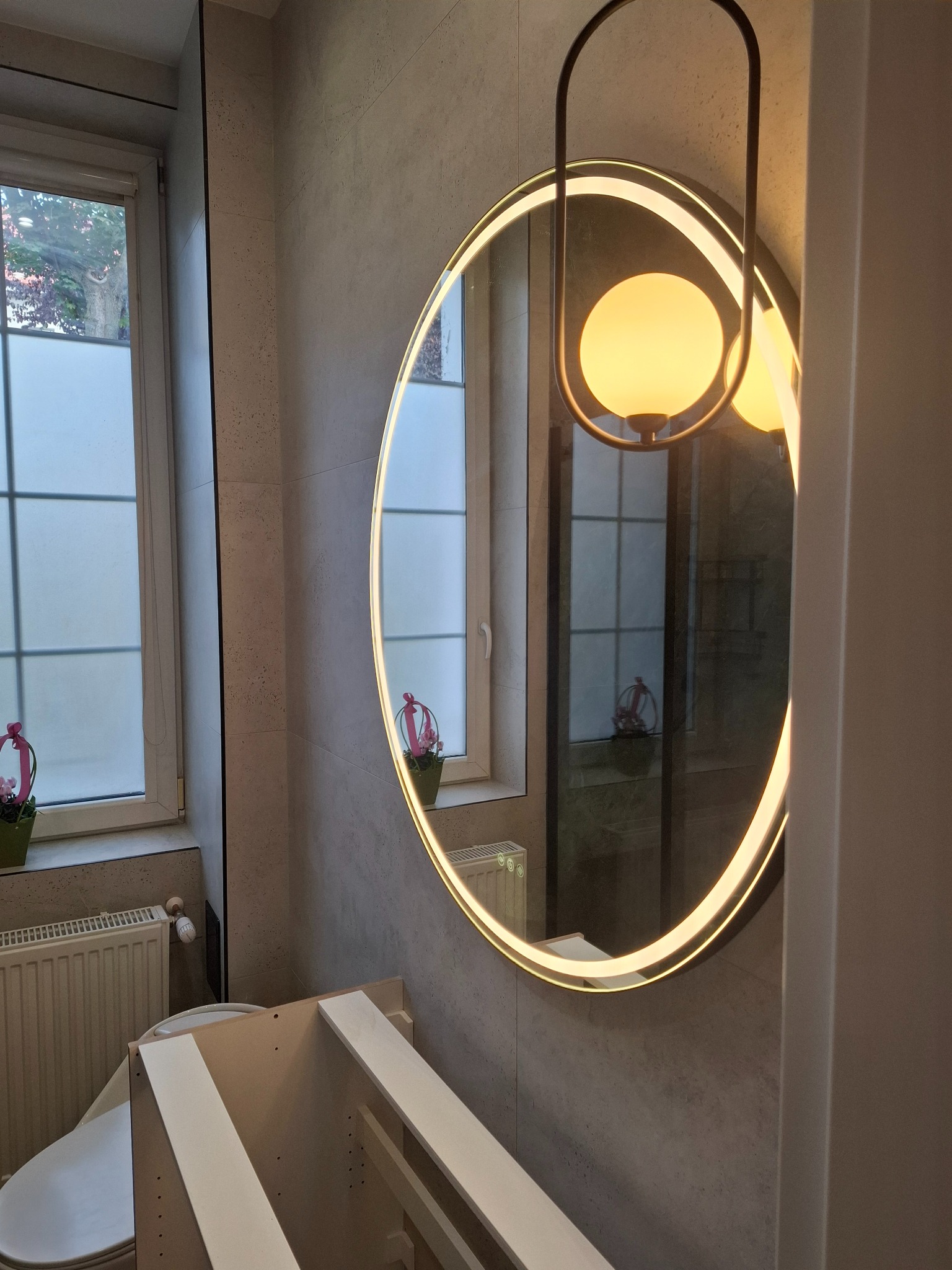 Okrągłe lustro z podświetleniem LED w łazience z szarą płytką. Widoczny fragment toalety i niedokończona szafka pod umywalkę. W tle okno z roletą.