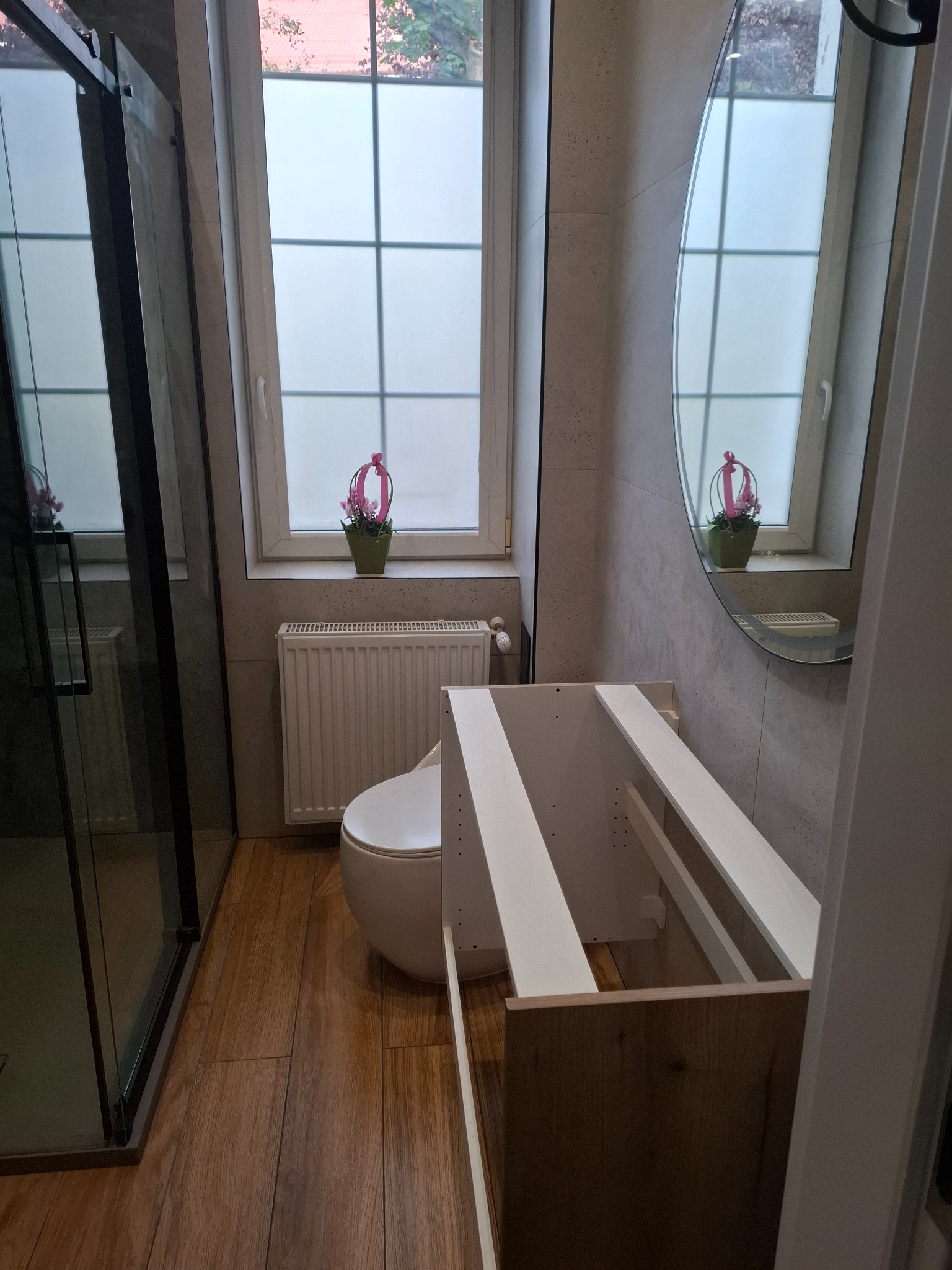 Nowoczesna łazienka w trakcie montażu: kabina prysznicowa, toaleta, grzejnik i szafka pod umywalkę z drewnianym frontem. Okrągłe lustro odbija kwiat w doniczce na parapecie.