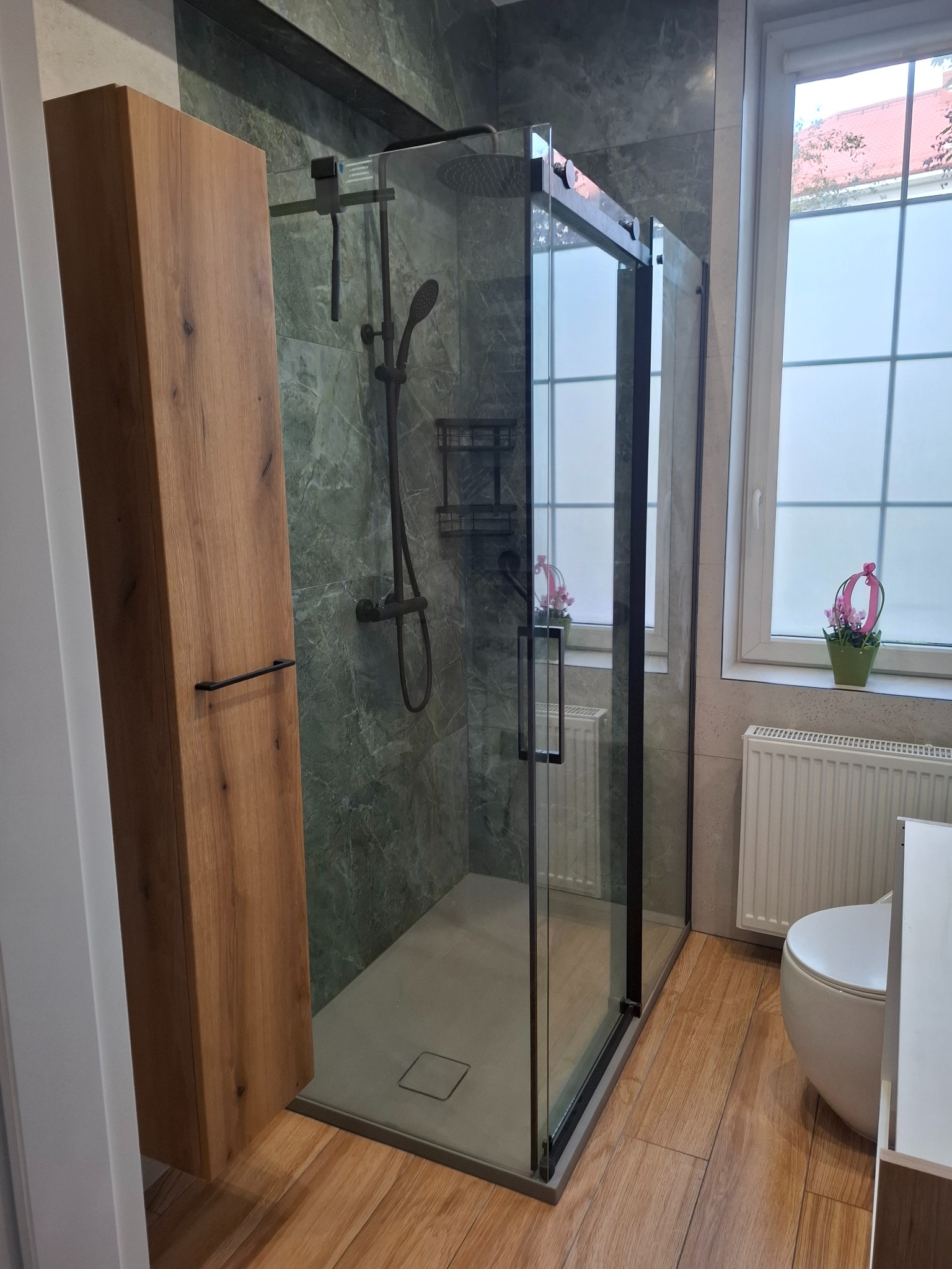 Nowoczesna kabina prysznicowa z czarnymi profilami, drewniana szafka łazienkowa i marmurowe płytki w odcieniach szarości. Widok na okno z kwiatami.