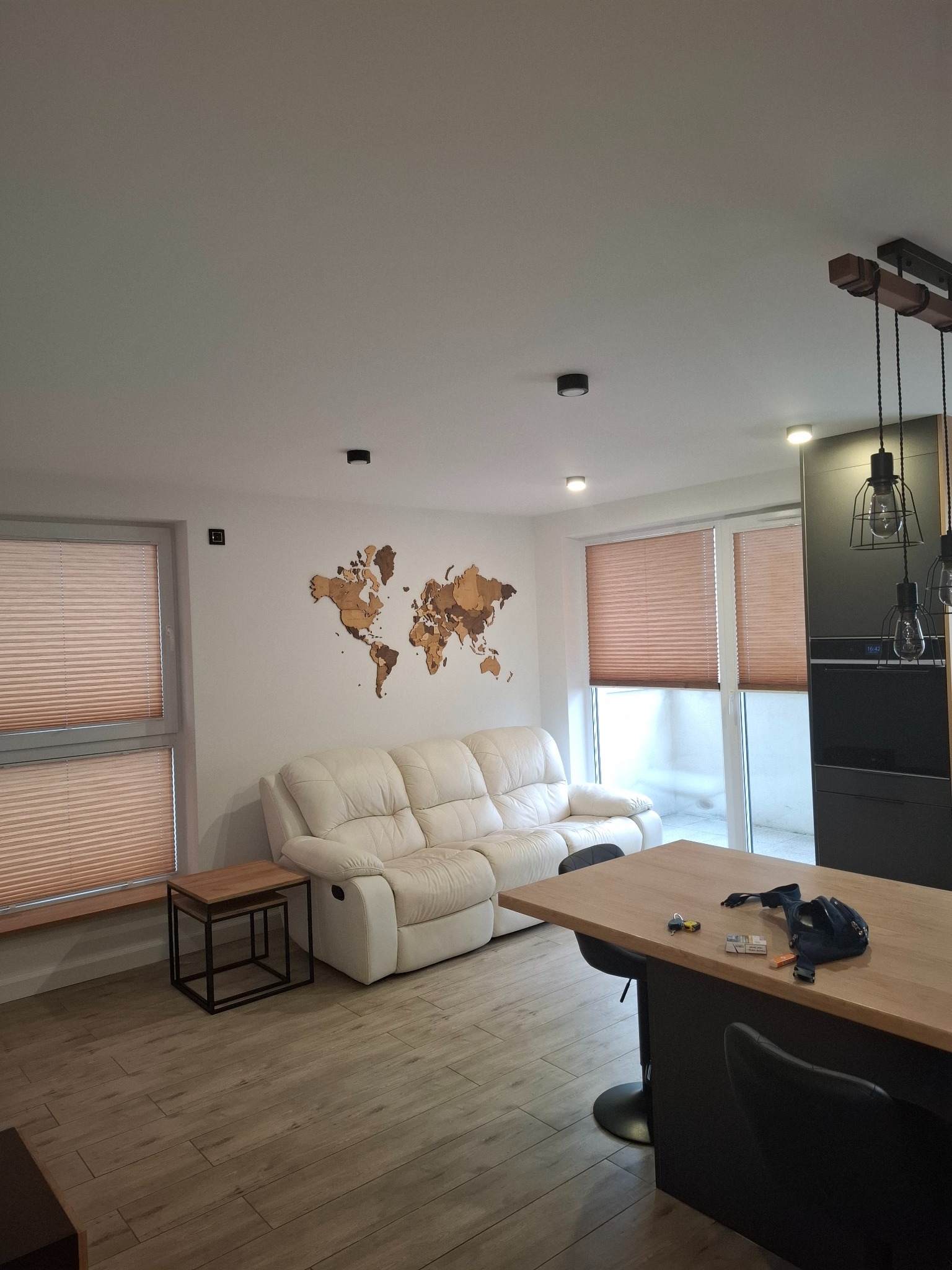 Jasne wnętrze: drewniana mapa świata na ścianie nad białą sofą, drewniany stół z czarnymi krzesłami barowymi, okna z żaluzjami i nowoczesne oświetlenie.