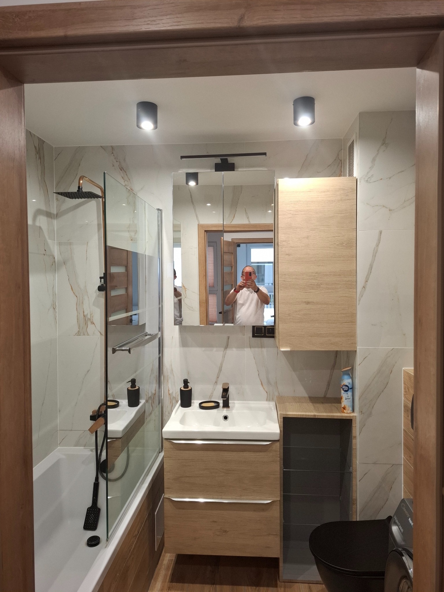 Nowoczesna łazienka z kabiną prysznicową typu walk-in, umywalką z szafką z drewna i czarną toaletą. Na ścianach płytki imitujące marmur. Widoczne oświetlenie punktowe.