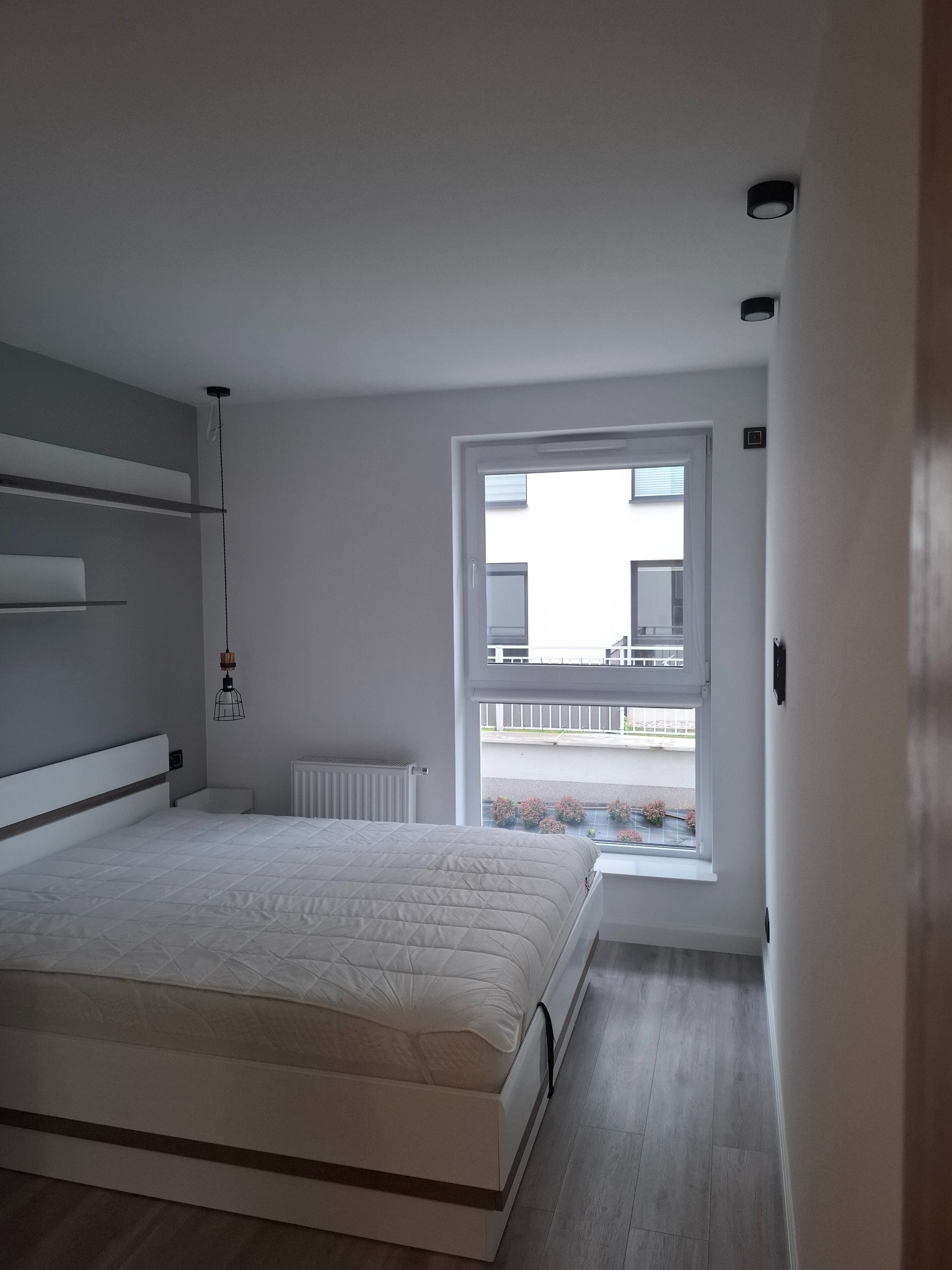 Jasna sypialnia z białym łóżkiem, drewnianą podłogą i oknem z widokiem na zieleń. Minimalistyczny design, lampa wisząca i półki na ścianie. Nowoczesne oświetlenie sufitowe.