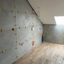 Łazienka w trakcie remontu na poddaszu: szare płytki ścienne z żółtymi krzyżykami dystansowymi, drewnopodobna podłoga, widoczne instalacje, fragment brodzika.