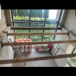 Wnętrze w trakcie ocieplania poddasza: widoczne drewniane belki konstrukcyjne, rolki wełny mineralnej Ursa, drabina i narzędzia na tle okna z widokiem na zieleń.