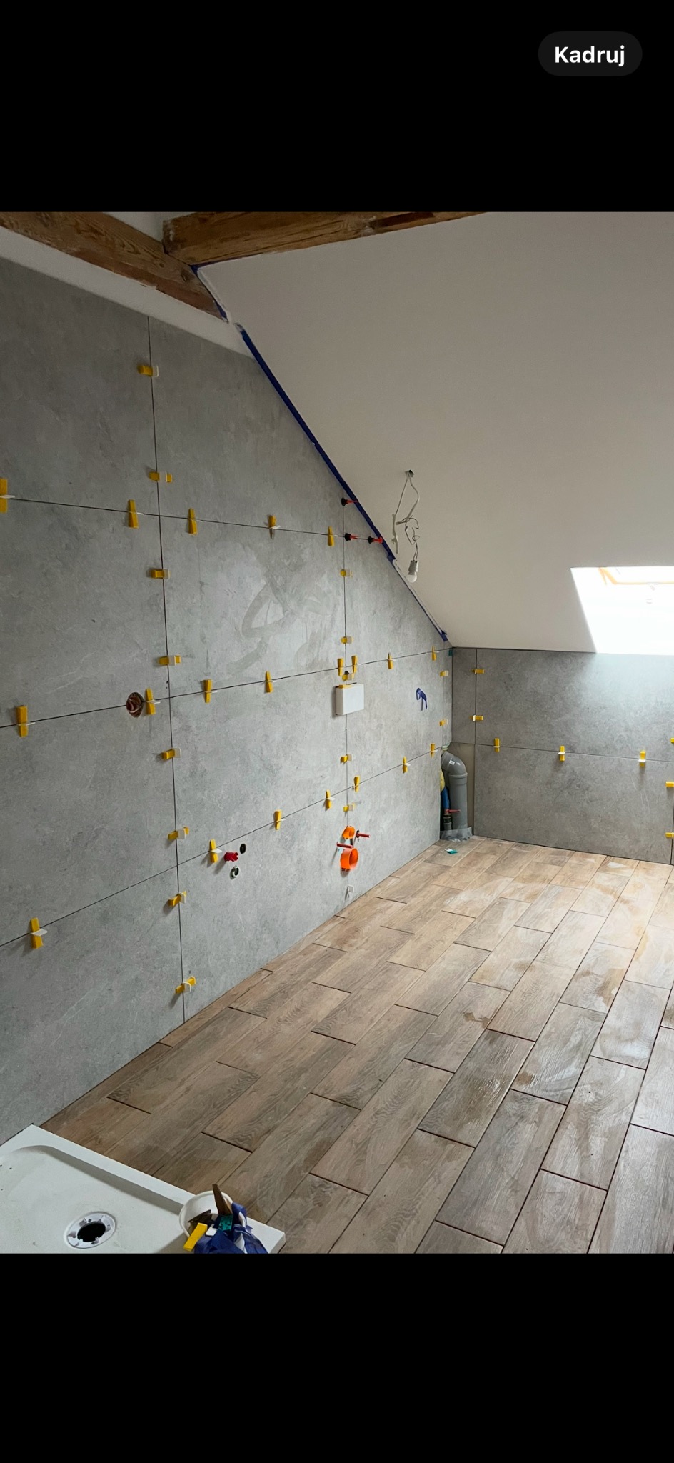 Łazienka w trakcie remontu na poddaszu: szare płytki ścienne z żółtymi krzyżykami dystansowymi, drewnopodobna podłoga, widoczne instalacje, fragment brodzika.