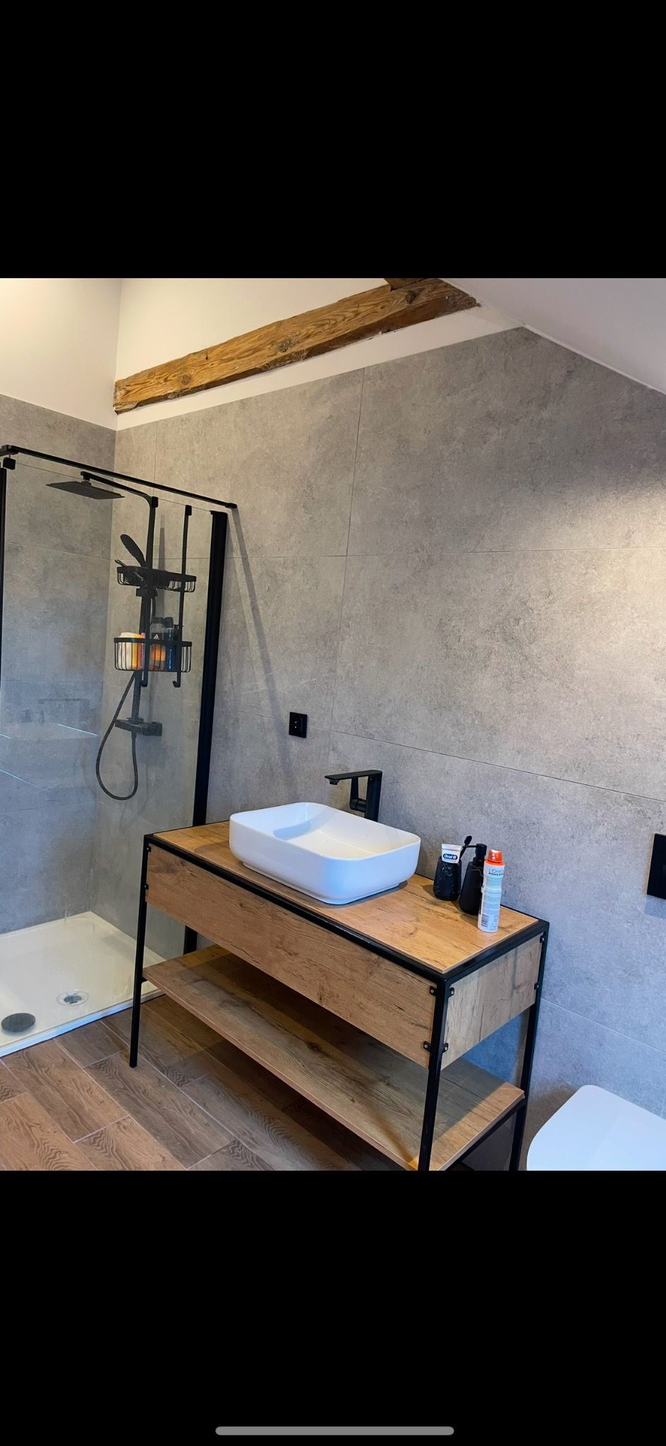 Nowoczesna łazienka z kabiną prysznicową typu walk-in, umywalką nablatową na drewnianej szafce z czarnymi metalowymi elementami i szarą płytką na ścianach, z drewnianą belką stropową.