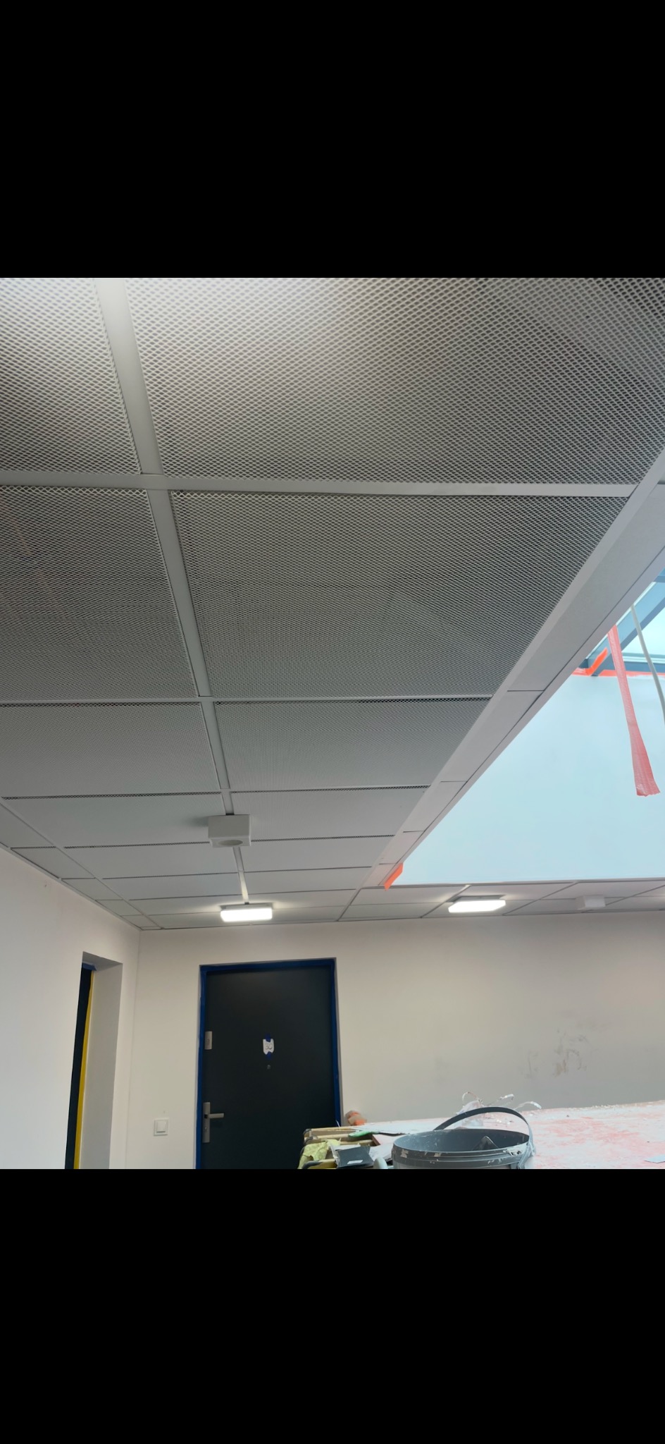 Sufit podwieszany rastrowy w biurze, widoczne lampy, drzwi i ślady prac remontowych na ścianie.
