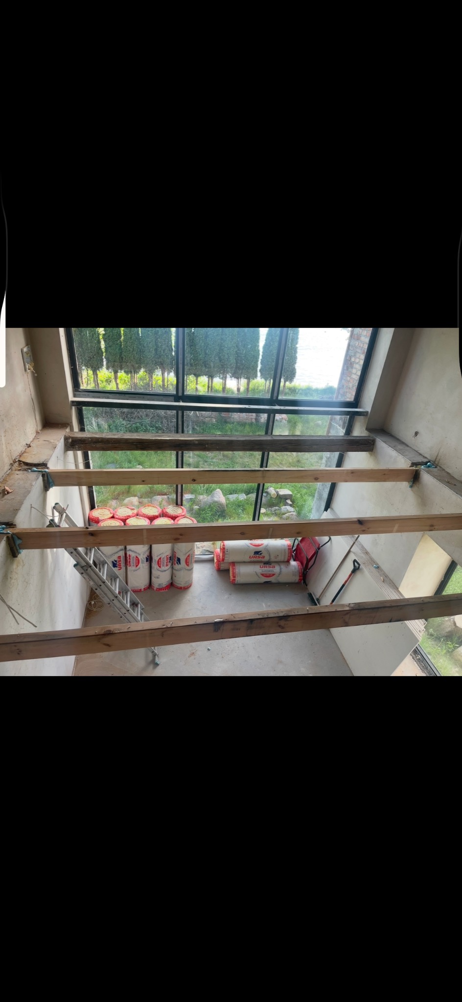 Wnętrze w trakcie ocieplania poddasza: widoczne drewniane belki konstrukcyjne, rolki wełny mineralnej Ursa, drabina i narzędzia na tle okna z widokiem na zieleń.