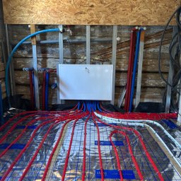 AquaTerm Ksawery Haduch - Instalacja ogrzewania podłogowego w trakcie montażu: czerwone rury układane na srebrnej folii izolacyjnej, widoczne niebieskie i czerwone przewody przy ścianie.