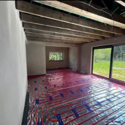 Widok na ułożone, czerwone rury ogrzewania podłogowego na srebrnej folii izolacyjnej w surowym wnętrzu z oknem i belkami stropowymi.