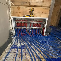 AquaTerm Ksawery Haduch - Rozdzielacz ogrzewania podłogowego z czerwono-niebieskimi rurami na izolacji, w tle drewniana ściana i wkrętarka DeWalt na szafce rozdzielczej.