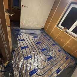 AquaTerm Ksawery Haduch - Ułożone, niebieskie rury ogrzewania podłogowego na folii aluminiowej, widoczne elementy drewnianej konstrukcji poddasza i okno. Etap prac przed wylaniem posadzki.