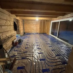 AquaTerm Ksawery Haduch - Instalacja ogrzewania podłogowego w surowym drewnianym wnętrzu. Widoczne rury grzewcze ułożone na folii, okno przesuwne i drewniana konstrukcja ścian.