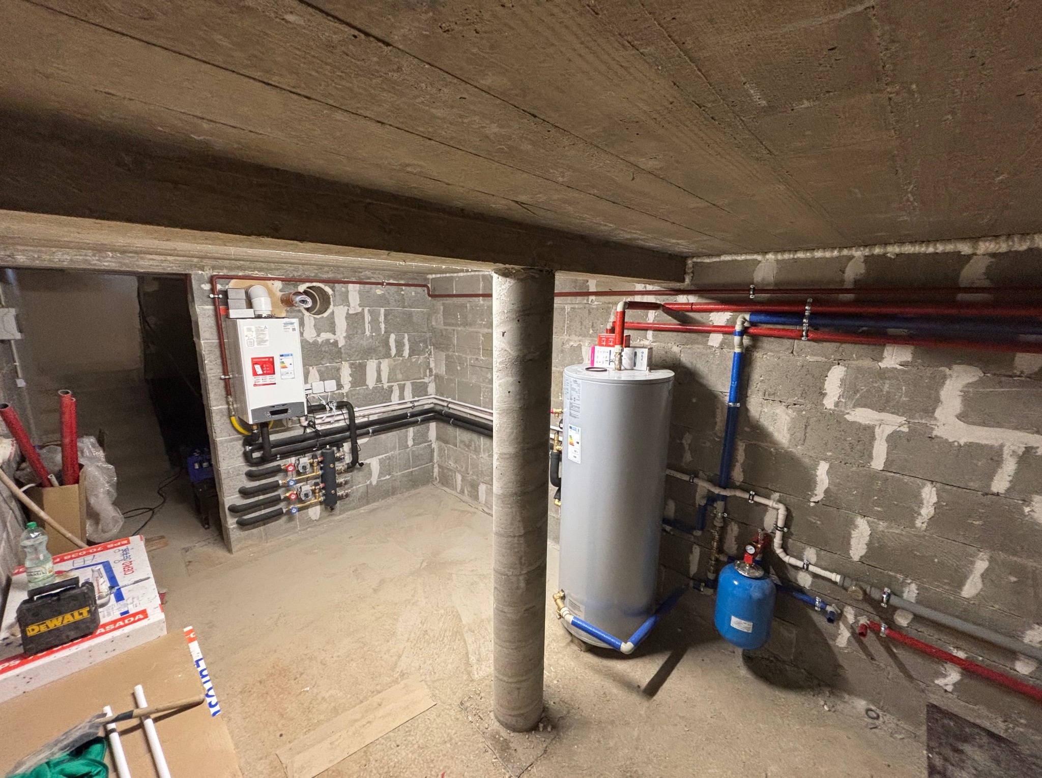 Szara piwnica z instalacją grzewczą: bojler, piec gazowy, rury czerwone i niebieskie. Betonowy sufit i ściany z bloków. Widoczne narzędzia i materiały budowlane.