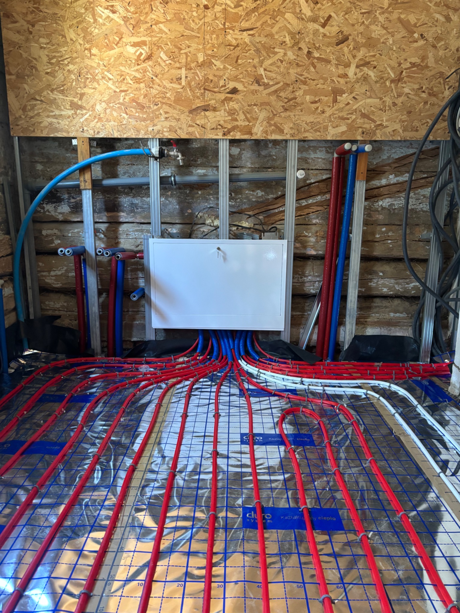 Instalacja ogrzewania podłogowego w trakcie montażu: czerwone rury układane na srebrnej folii izolacyjnej, widoczne niebieskie i czerwone przewody przy ścianie.