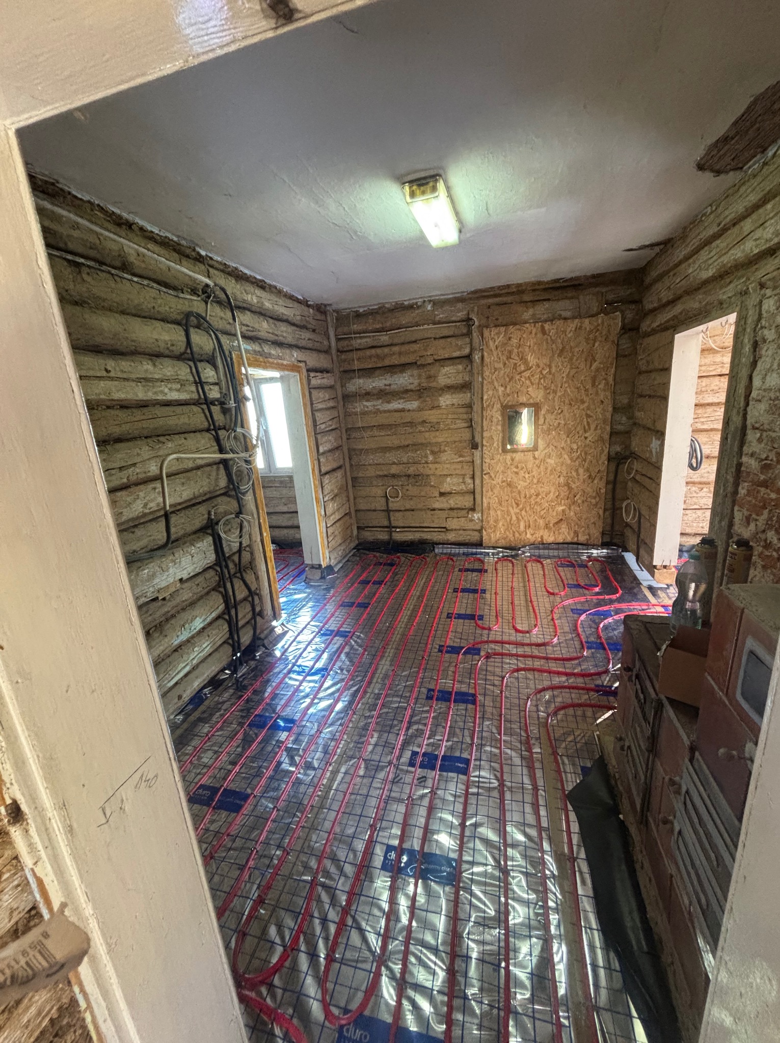 Instalacja ogrzewania podłogowego w drewnianym domu. Czerwone rurki ułożone na folii izolacyjnej, widoczne ściany z bali i instalacje elektryczne.