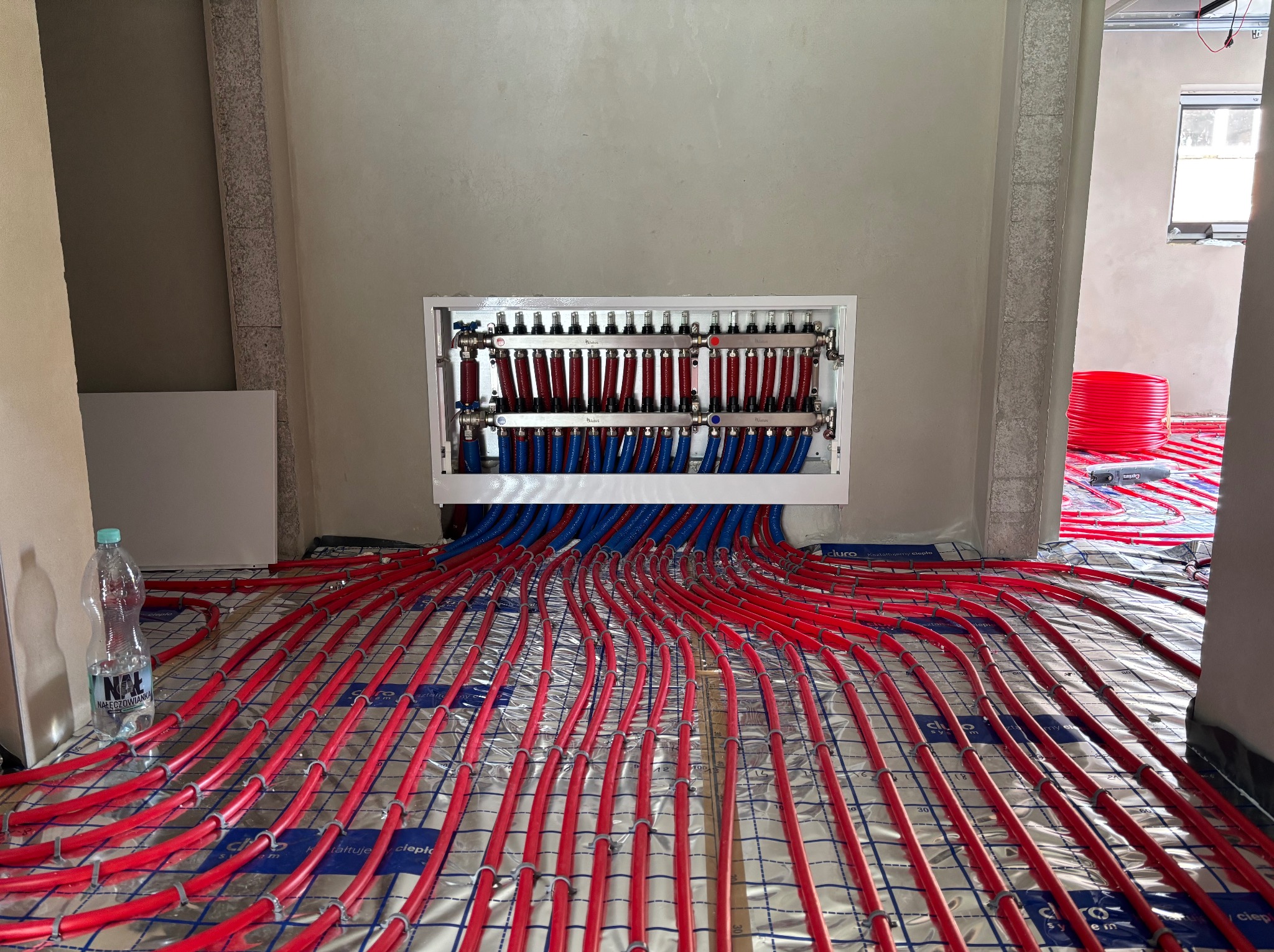 Rozłożona instalacja ogrzewania podłogowego z czerwonymi rurami na folii, widoczny rozdzielacz i butelka wody. Surowe ściany w tle, okno w oddali.