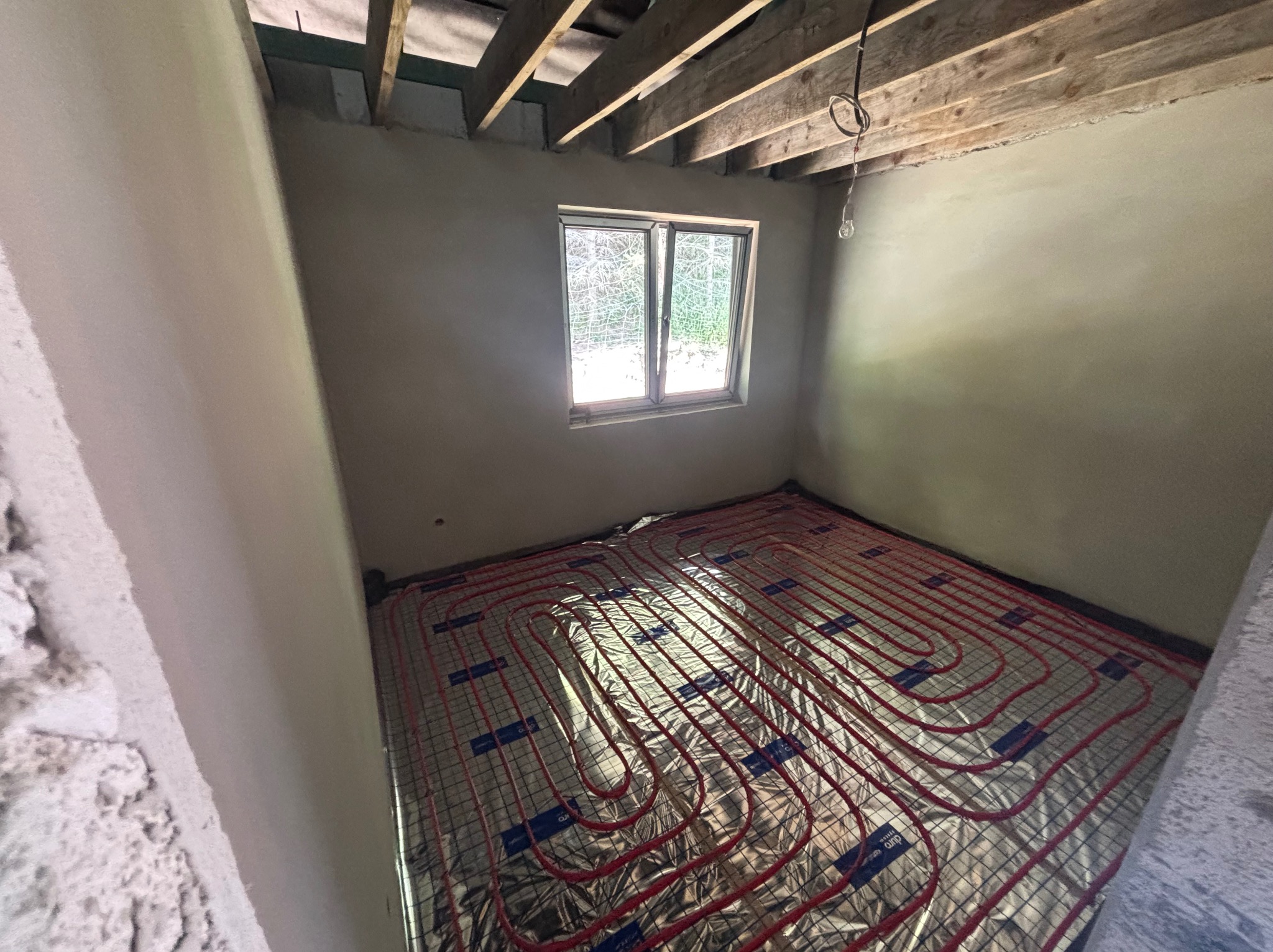 Ułożona instalacja ogrzewania podłogowego z czerwonymi rurkami na srebrnej macie w surowym pomieszczeniu z oknem i drewnianym stropem, widok z góry.