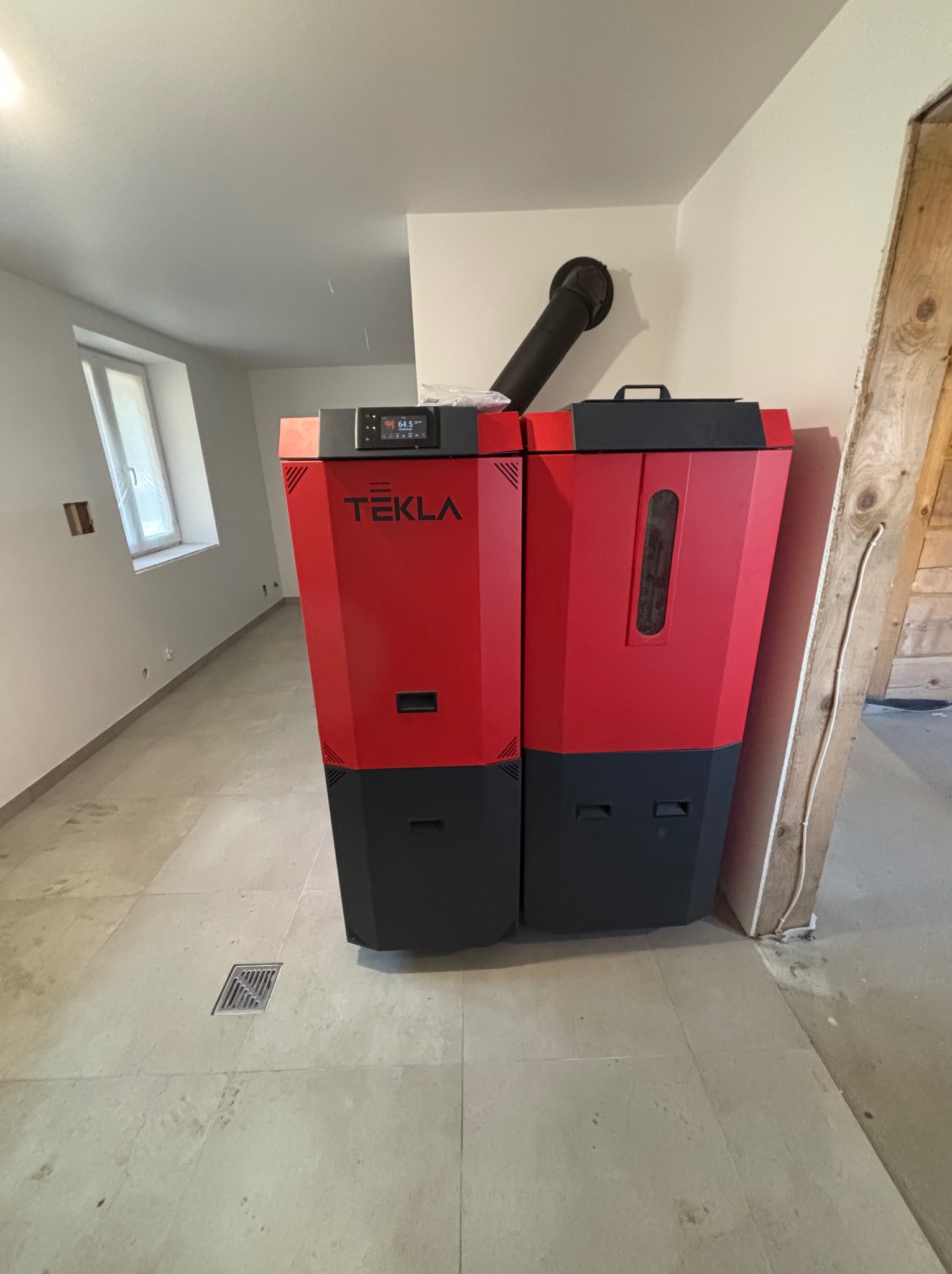 Nowoczesny piec CO marki Tekla, w kolorze czerwono-czarnym, z podłączonym kominem, zainstalowany w jasnym pomieszczeniu z oknem i drewnianą konstrukcją.