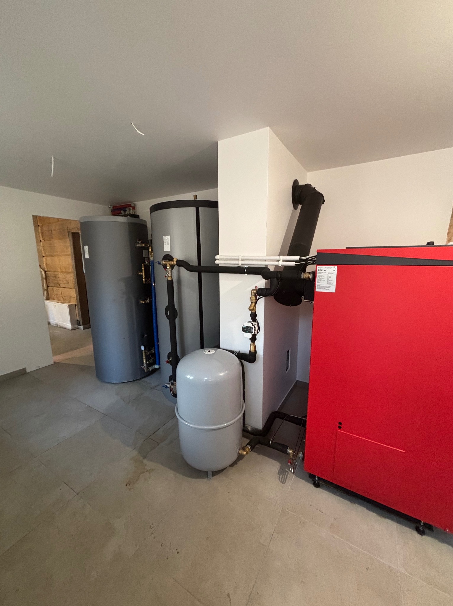 Zestaw kotła grzewczego Tekla z zbiornikami akumulacyjnymi w kotłowni, rury instalacji CO, czarny komin, szara podłoga, widok z boku na czerwony korpus kotła.