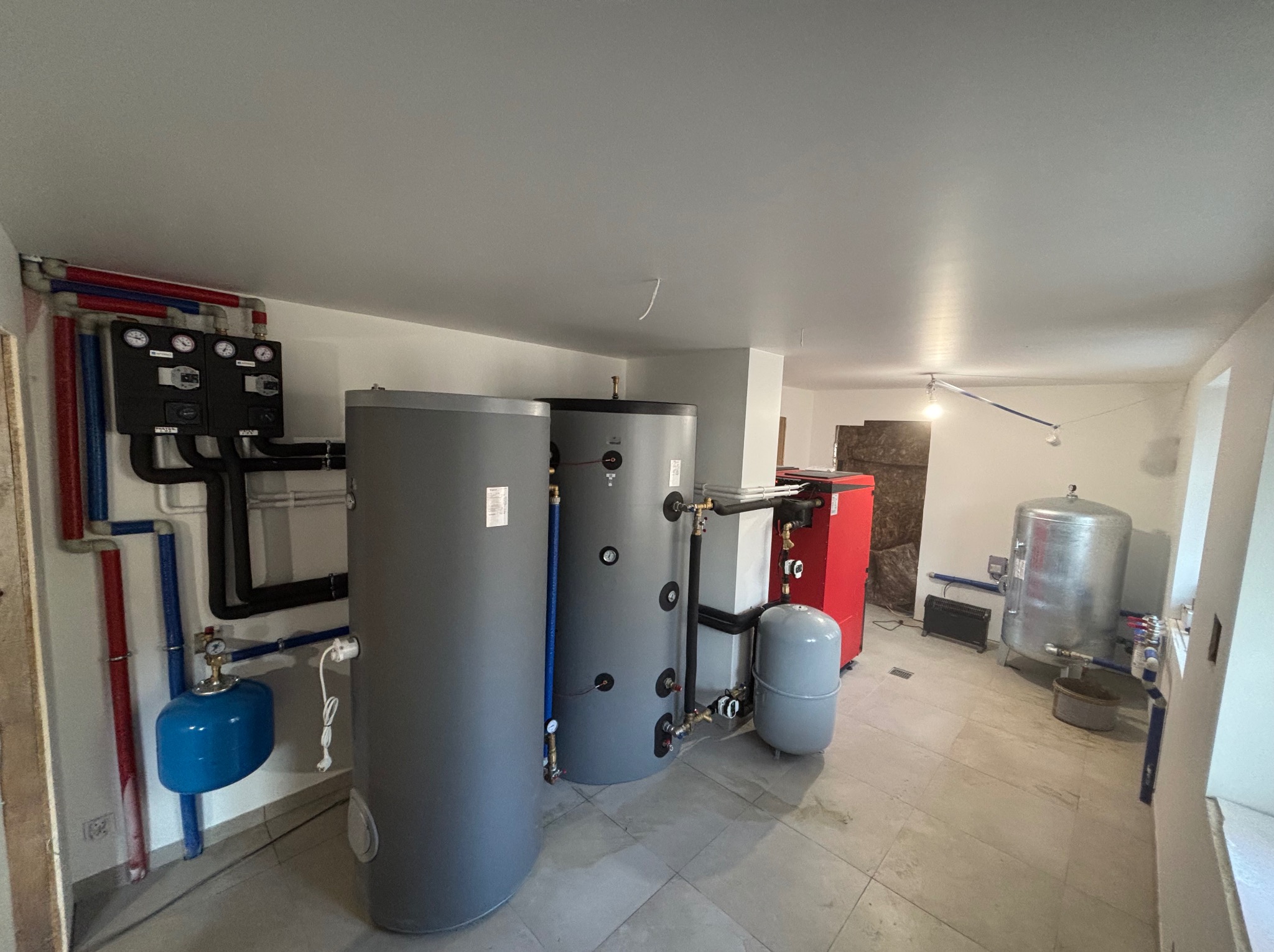 Kompleksowa kotłownia: dwa szare zbiorniki buforowe, czerwony piec CO, czarna automatyka, instalacja z rur w kolorach czerwonym i niebieskim, szare naczynie przeponowe.