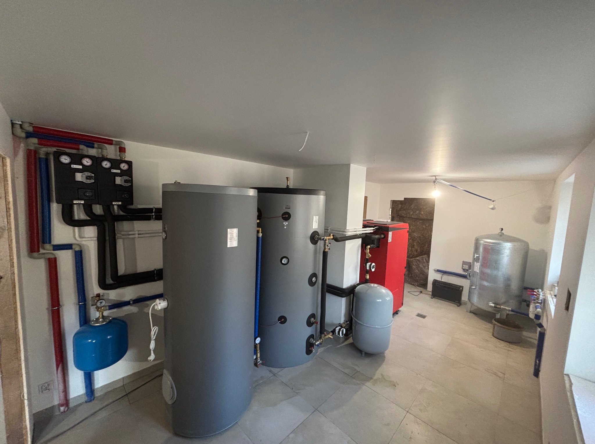 Zestaw urządzeń grzewczych w kotłowni: dwa szare zasobniki, czerwony piec CO, zbiornik wyrównawczy, czarna rozdzielnica z manometrami, instalacja rurowa.