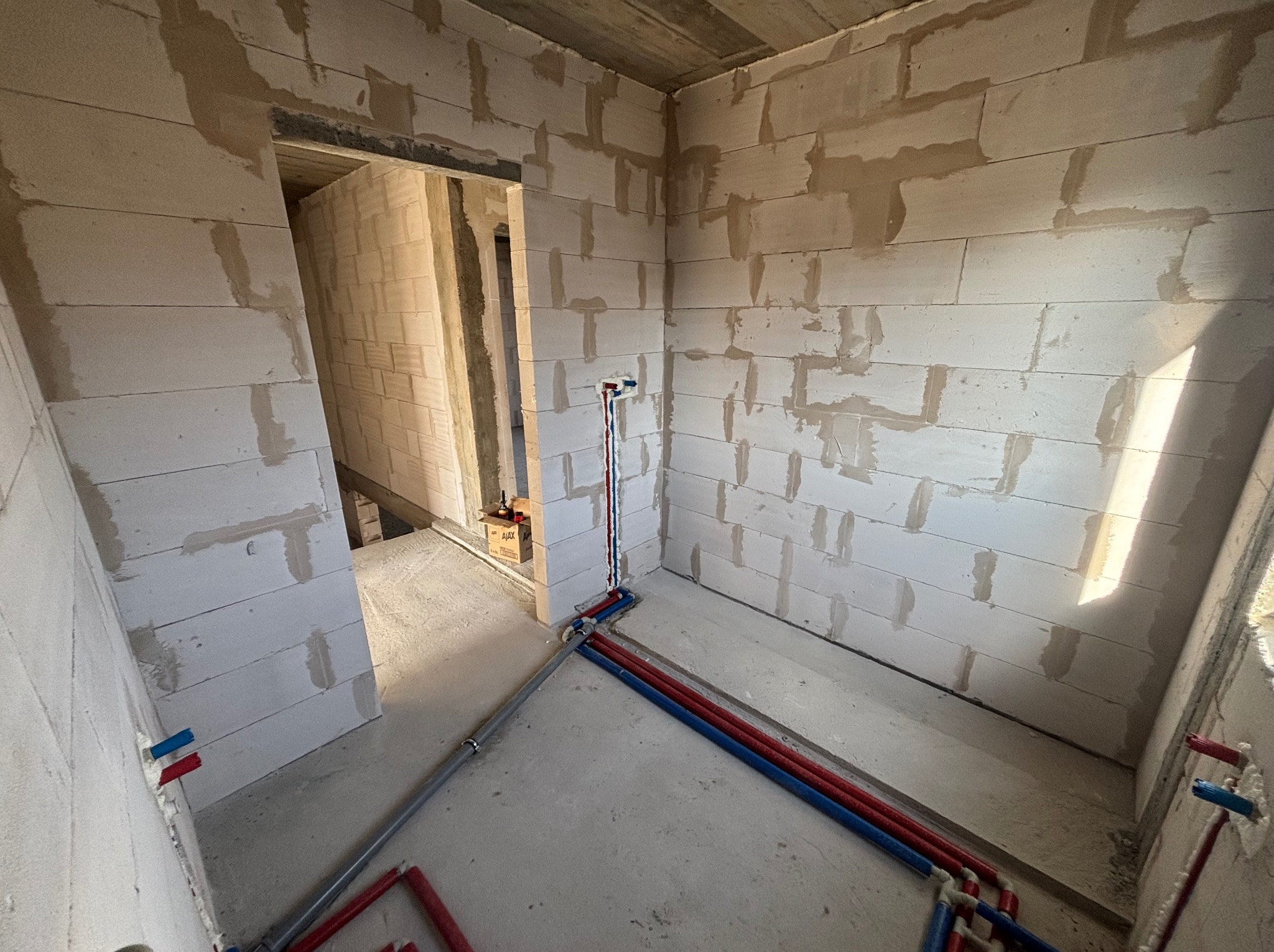 Wnętrze w surowym stanie z widocznymi rurami instalacji wodnej (czerwone i niebieskie) wychodzącymi ze ściany i rozprowadzonymi po podłodze. Ściany z białego betonu komórkowego.