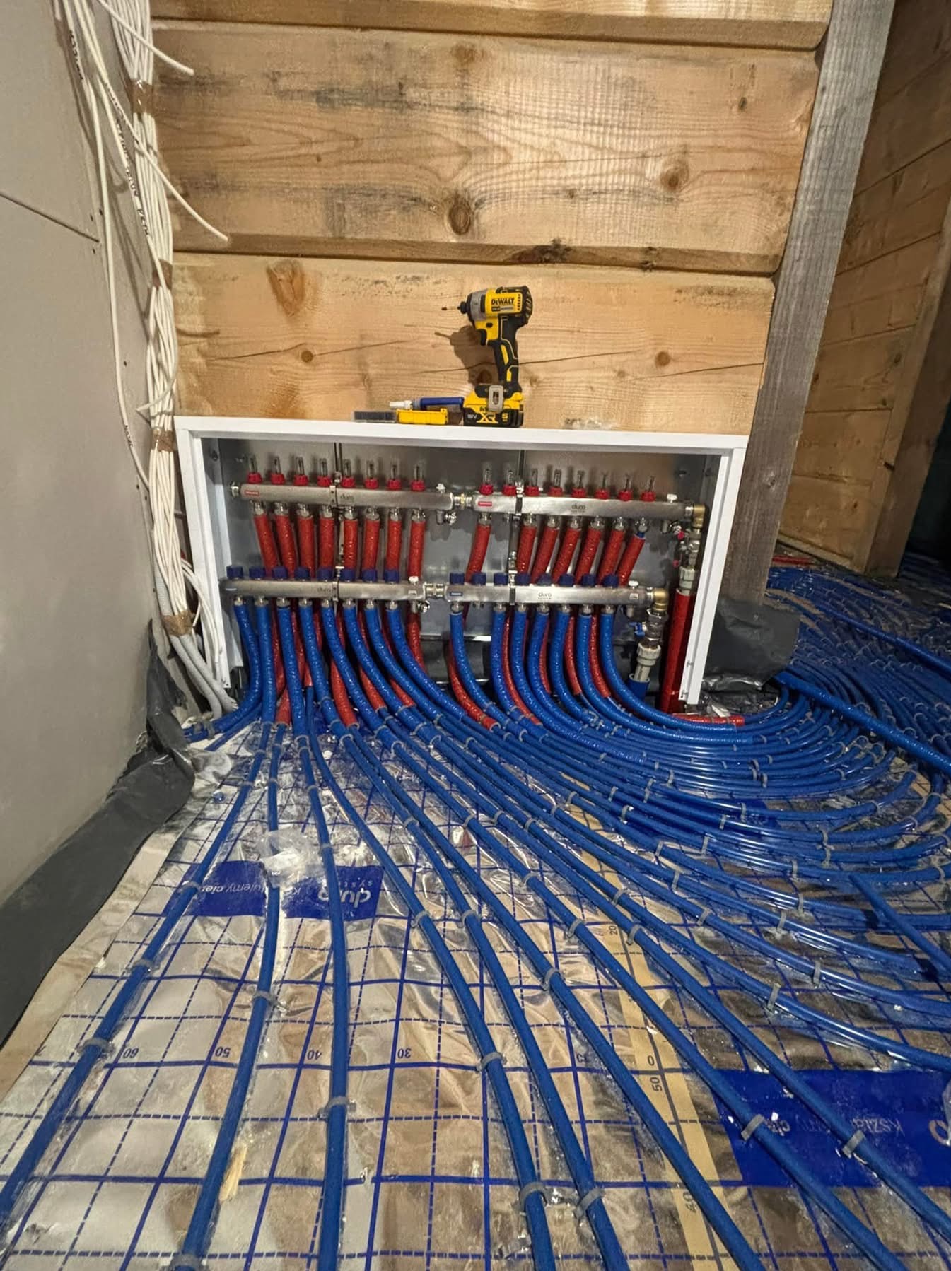 Rozdzielacz ogrzewania podłogowego z czerwono-niebieskimi rurami na izolacji, w tle drewniana ściana i wkrętarka DeWalt na szafce rozdzielczej.