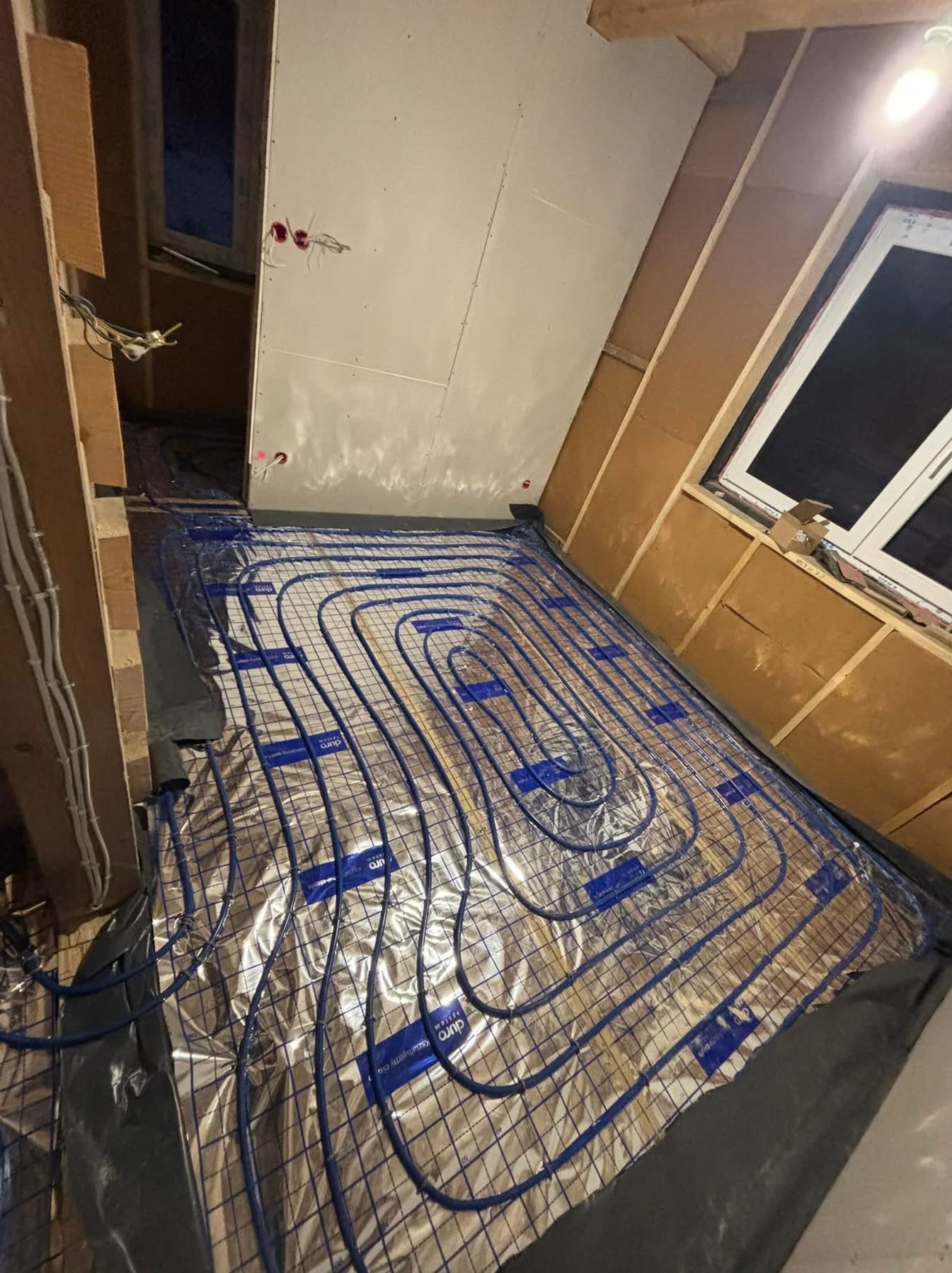 Ułożone, niebieskie rury ogrzewania podłogowego na folii aluminiowej, widoczne elementy drewnianej konstrukcji poddasza i okno. Etap prac przed wylaniem posadzki.