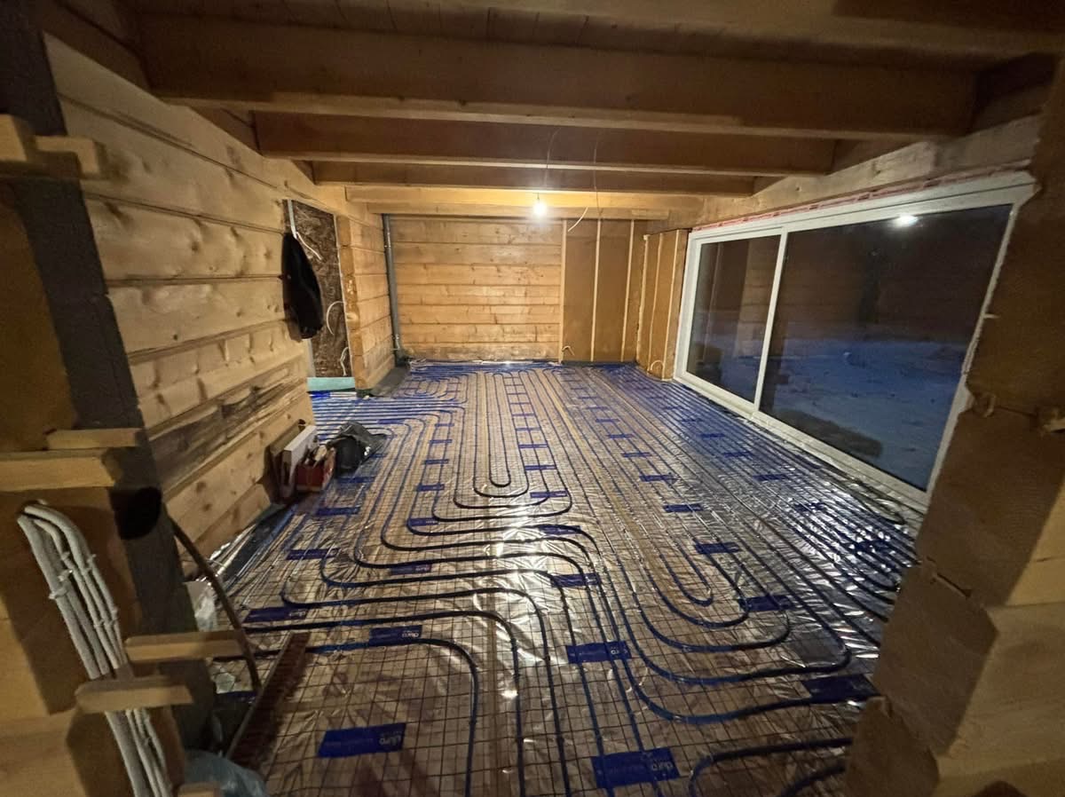 Instalacja ogrzewania podłogowego w surowym drewnianym wnętrzu. Widoczne rury grzewcze ułożone na folii, okno przesuwne i drewniana konstrukcja ścian.